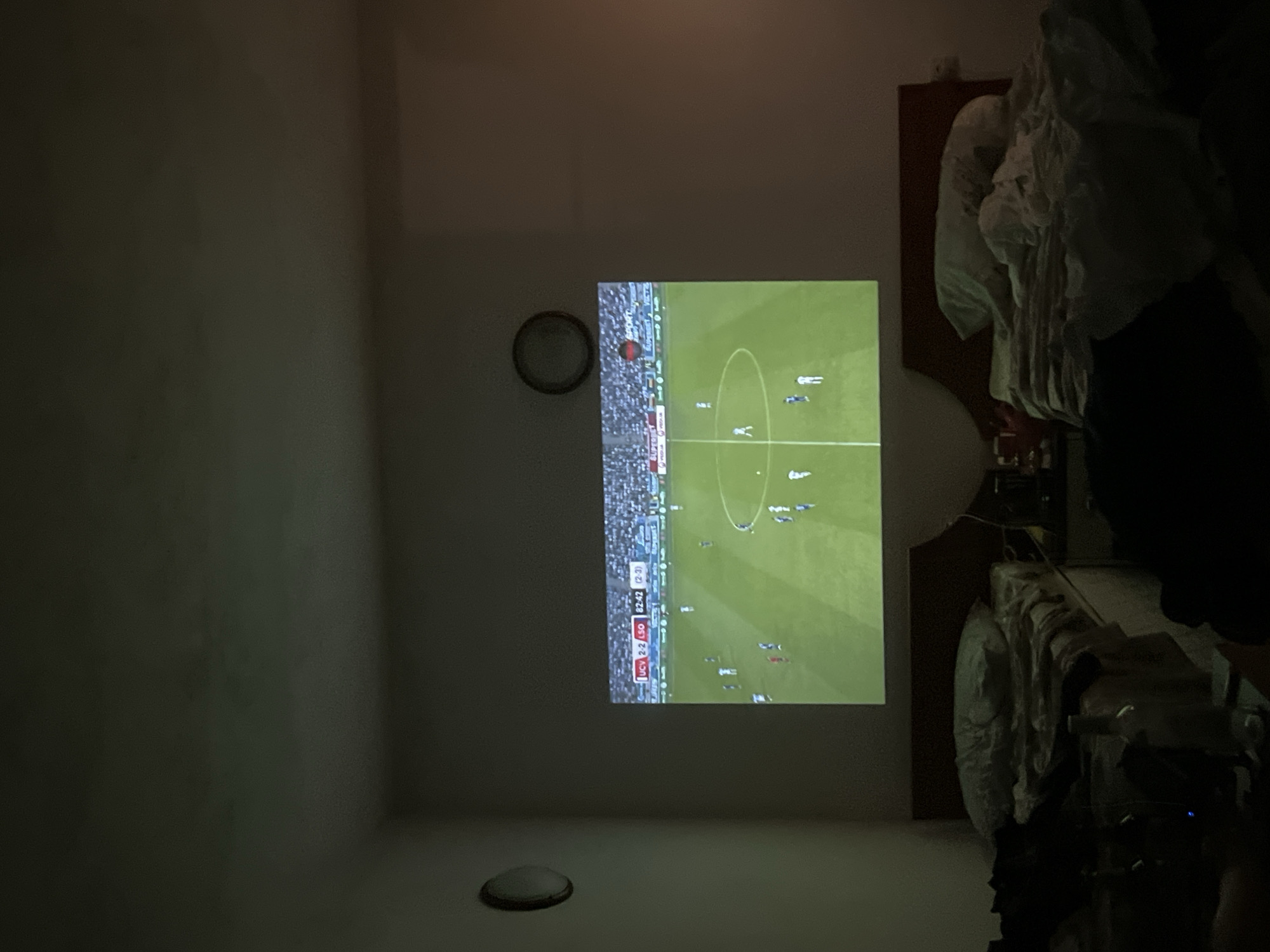 This is a medium shot, taken from a slightly elevated angle, of a bedroom at night in Obzor, Bulgaria. The room is dark, illuminated primarily by a projector displaying a soccer match on the wall. The projector displays a soccer field with players in white and blue uniforms, and a scoreboard at the top left shows "UCV 2-2 LSO 82:42 (2-3)". Two beds with rumpled white bedding and pillows are visible in the foreground, positioned side-by-side. Various objects, including bottles and a lamp, are scattered on the nightstand between the beds and on the floor. A circular light fixture is mounted on the wall to the left of the projector screen, and a clock is visible on the wall to the right. The overall impression is one of a casual, at-home viewing experience.
