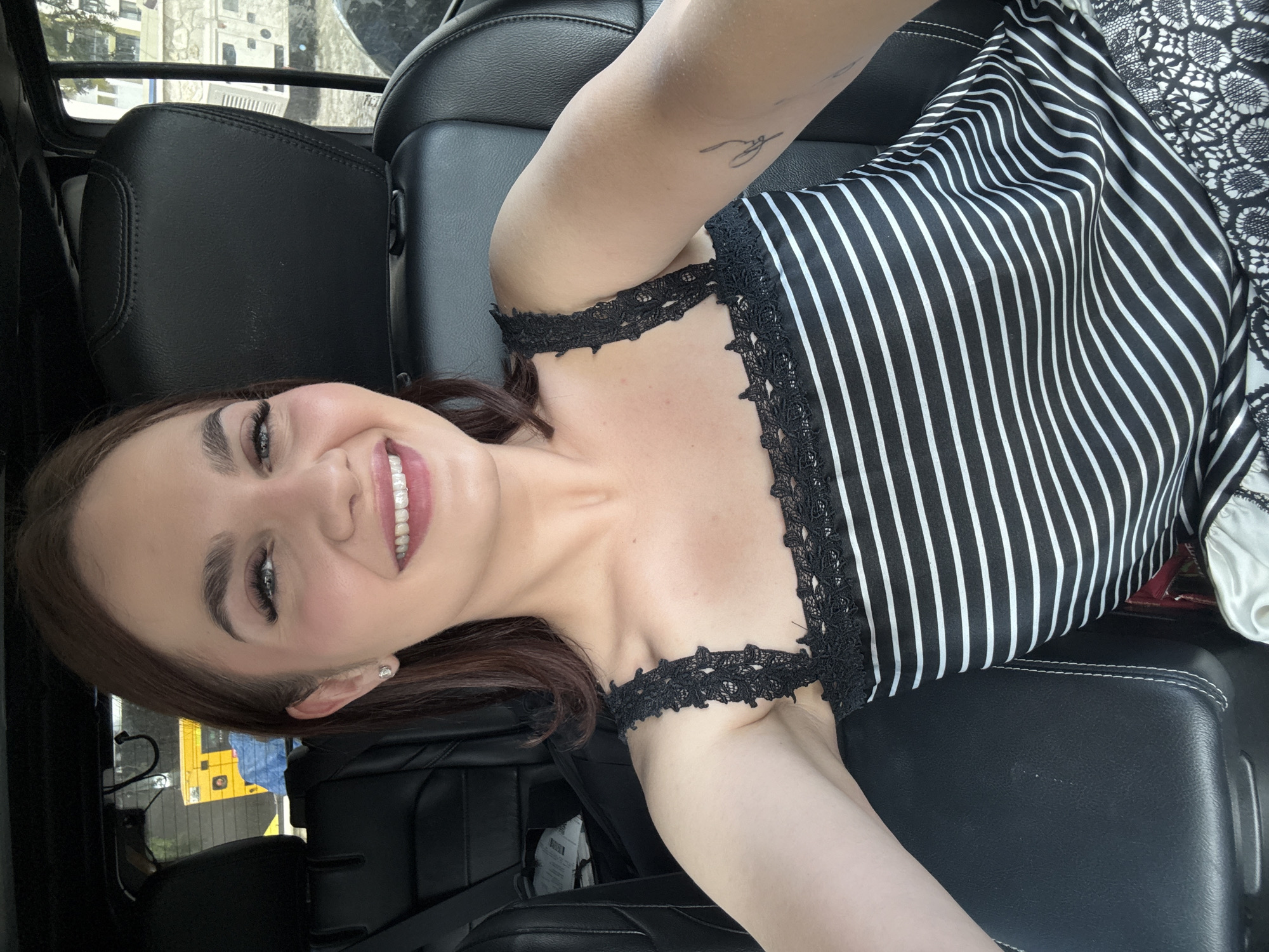 A young woman with brown hair is smiling and appears to be taking a selfie while seated in a car. She is wearing a black and white striped top with black lace straps. Her left arm is visible, with a small tattoo on the inner part of her upper arm. The car's interior is predominantly black, with leather seats. A glimpse of the outside environment is visible through the car window, revealing a cityscape with buildings and a yellow school bus. This cannot be used to determine the location in Sofia, Bulgaria.