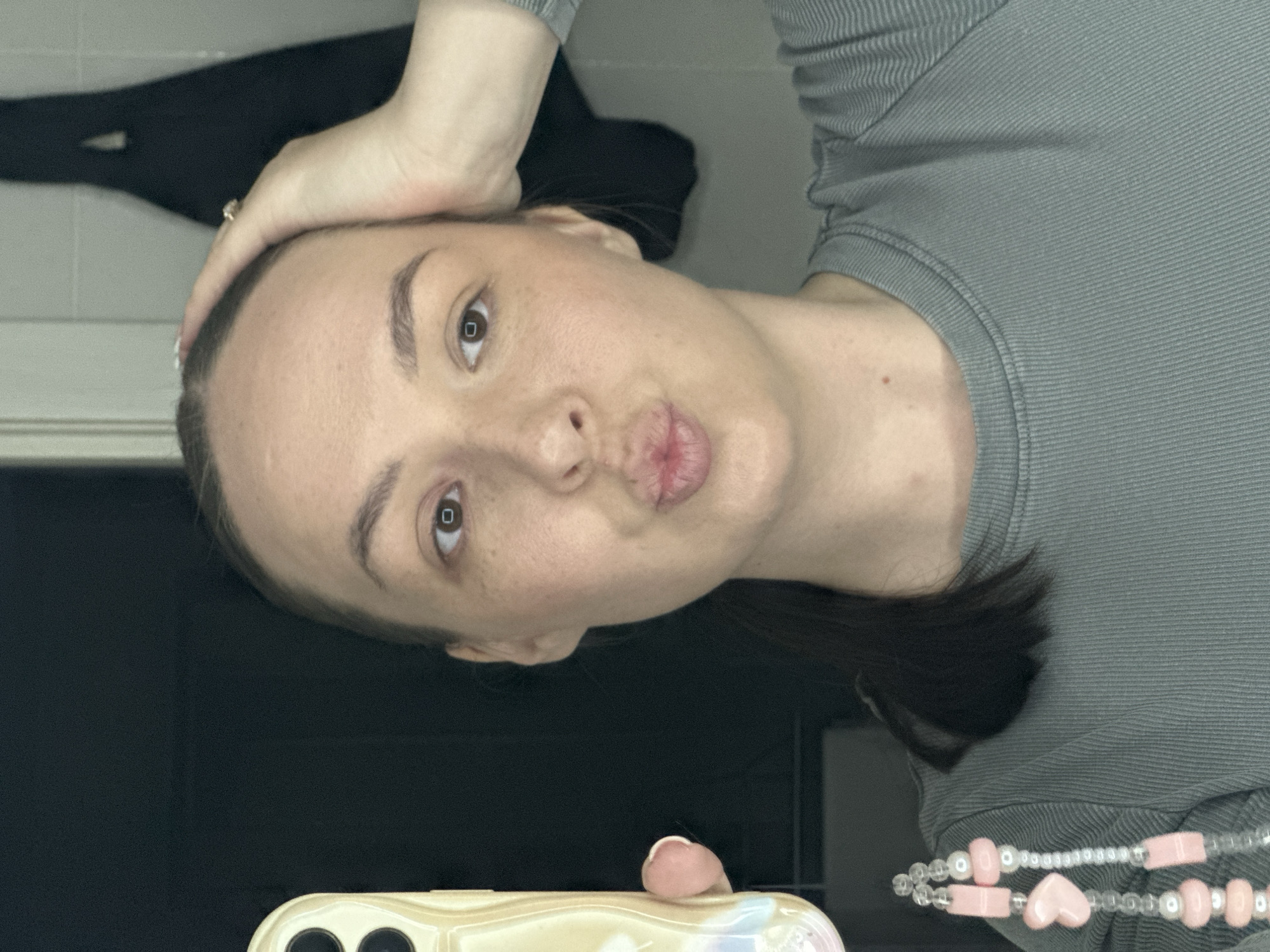 A woman is posing for a selfie. She has fair skin, brown eyes, and her hair is pulled back. She is wearing a gray ribbed shirt and a pink and white beaded necklace. Her left hand is on her head, and she is making a kissing face at the camera. The phone she is using to take the picture is visible at the bottom of the frame. The background is dark, with a hint of a white wall. There is a black garment visible in the background as well. There's no evidence to suggest the photo was taken in Chaikovsky, Russia.