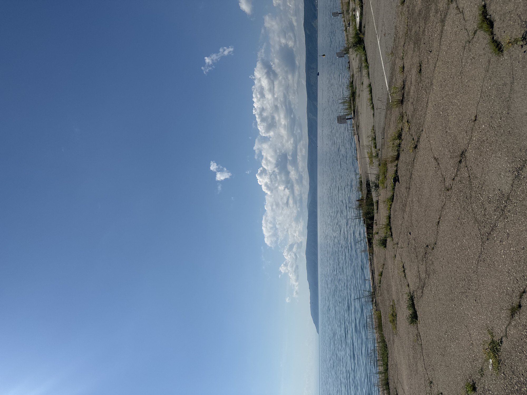 An outdoor, vertical, eye-level, medium shot captures a bright, sunny day at the shores of Lake Baikal in Listvyanka, Russia. The foreground is dominated by a cracked, grey asphalt surface, sparsely populated with weeds and dry grass. To the left, the vast, tranquil blue waters of Lake Baikal stretch out towards a distant, tree-covered mountain range on the horizon. The sky above is a clear, vibrant blue, with a few scattered white clouds, some appearing wispy and others more voluminous. In the middle ground, a white line is visible on the asphalt, suggesting a parking lot or road. Several white signs on posts are scattered near the waterline. A buoy with an orange top is visible in the water, and a dark boat is further out. The overall impression is one of natural beauty combined with a somewhat neglected man-made infrastructure.
