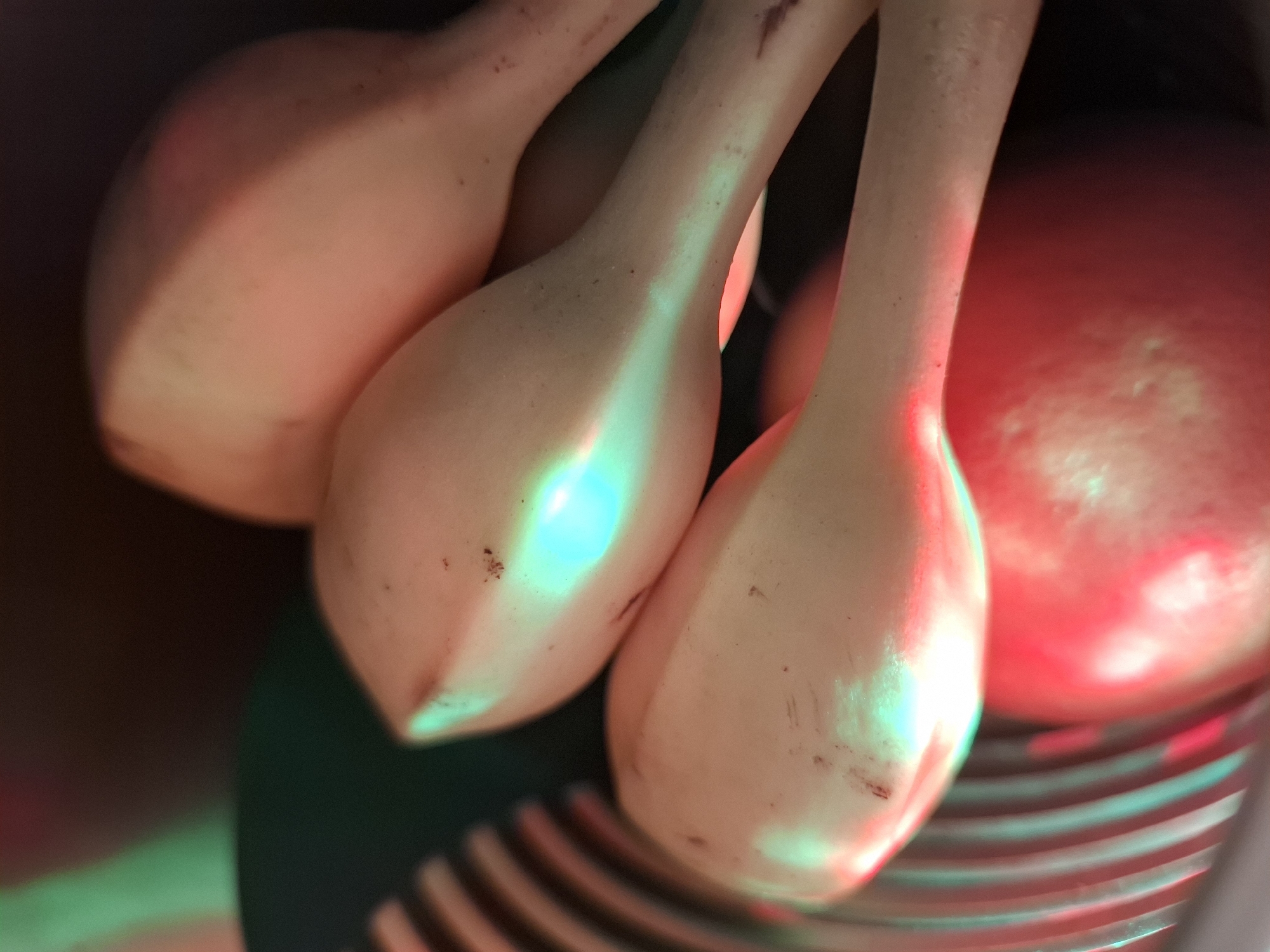The image shows a close-up, low-angle view of what appear to be several uncooked bananas. The bananas are pale yellow with some darker spots, and they are arranged in a cluster. The lighting in the scene is dramatic, with strong red and green colored lights creating reflections and highlights on the bananas. In the background, a red, spherical object is visible, possibly a tomato or an apple. The overall impression is one of an abstract still life, with the vibrant lighting playing a significant role in the composition. There are no people visible in the image. The location is described as Unknown, Unknown, which suggests it is not a specific, identifiable place within a city.