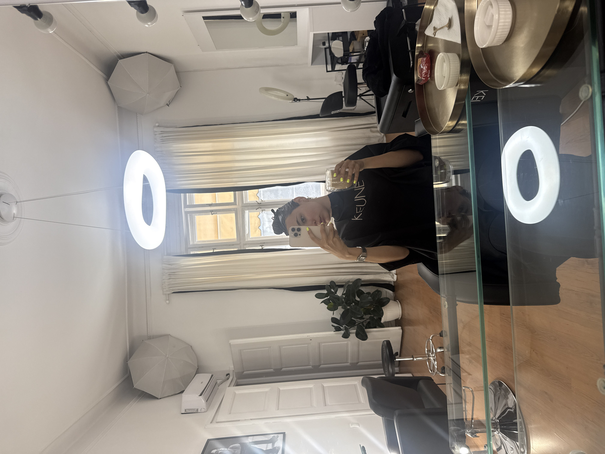A mirror selfie taken in a hair salon in Unknown, Unknown. A woman wearing a black t-shirt with "KEUNE" printed on it holds a smartphone and a glass of yellow liquid. The salon is equipped with a white ring light, a salon chair, a white umbrella-shaped softbox, and a plant. The window has white curtains and a closed blind. There is an air conditioning unit. The photo is taken in a mirror. A black coat is draped over a chair in the background. A glass shelf holds salon supplies.