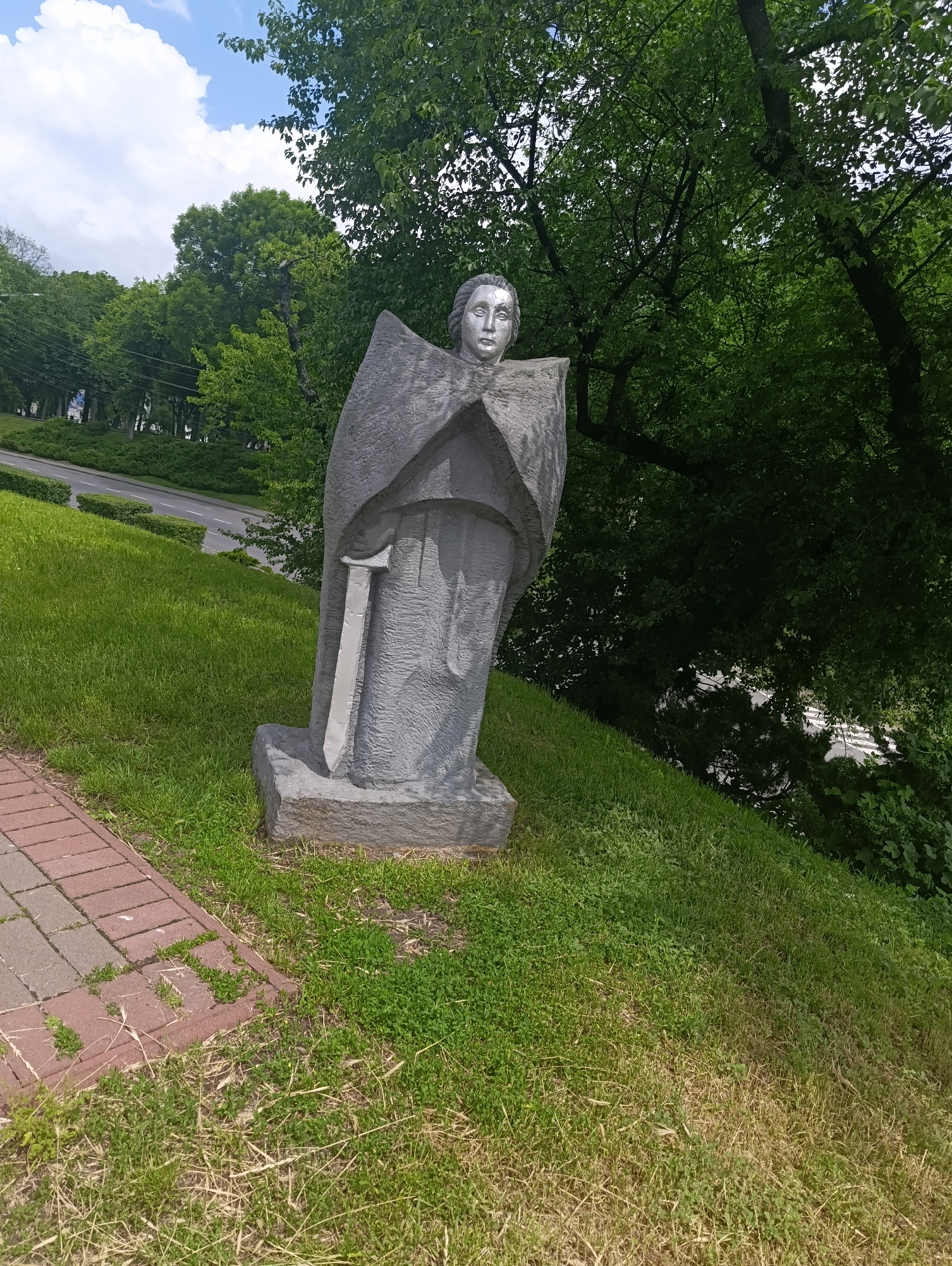 This is a photograph of a stone sculpture of a robed figure with a sword, situated outdoors on a grassy hillside. The sculpture is gray and abstract in style, with a stylized face and clothing that resembles wings or a cape. A sword is depicted within the folds of the robe. The sculpture stands on a rough stone base.
The hillside is covered in green grass, with some patches of dry grass and weeds. A brick pathway made of reddish-brown pavers is visible to the left of the sculpture, leading towards the foreground. In the background, a road curves uphill, bordered by neatly trimmed green hedges. Further back, mature trees with lush green foliage dominate the scene, with some branches extending into the sky. The sky is blue with scattered white clouds.
This sculpture is located in the city of Chernihiv, Ukraine, specifically on the Territory of the Church of the Assumption of the Blessed Virgin Mary (also known as the Spaso-Preobrazhensky Cathedral).