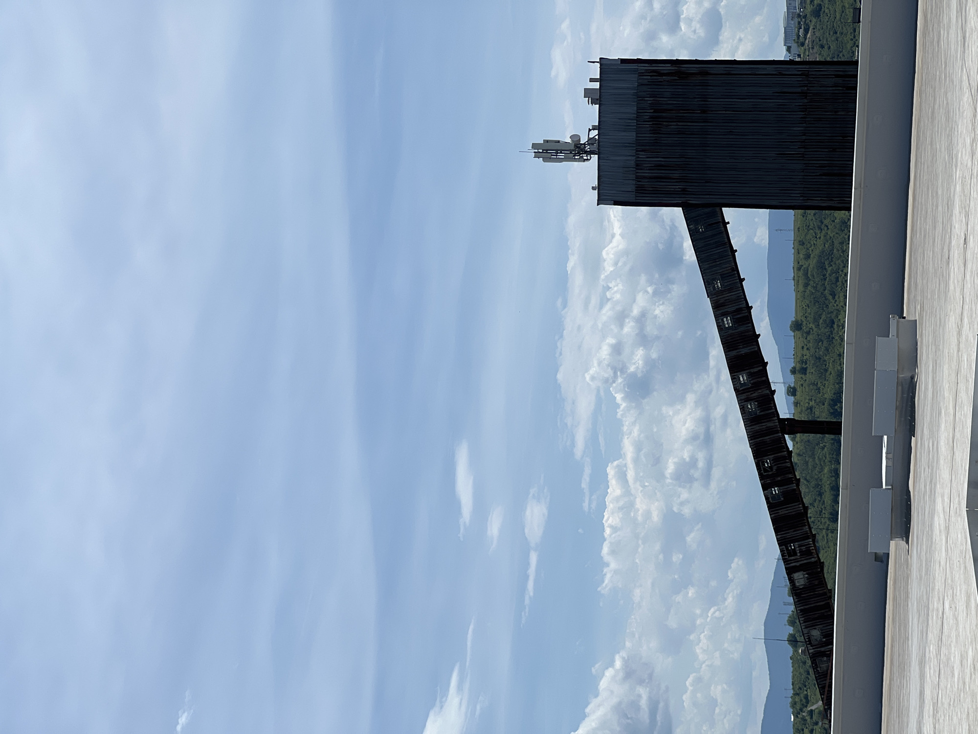 The image displays an outdoor scene dominated by industrial structures and a vast sky, under bright daytime conditions. The photograph appears to be rotated 90 degrees counter-clockwise, showing a large portion of blue sky and clouds on the left and top, and ground/structures on the right and bottom.
In the mid-ground, a prominent, tall industrial building constructed from dark, corrugated metal stands on what appears to be a large, flat, light-colored rooftop visible on the lower right. Mounted on top of this building is an array of communication antennae, dishes, and other equipment. Extending from the main building, sloping downwards and to the left, is a long, dark conveyor belt system or elevated walkway, featuring small, rectangular window-like openings along its side.
Beyond these structures, in the background, a range of green, tree-covered hills or mountains stretches across the horizon. The sky is a bright, clear blue, filled with various white clouds; some are thin and wispy, while others are large and puffy cumulus formations, particularly dense towards the bottom right of the sky (when viewing the image in its current rotated orientation).
No people are visible, and there is no explicit activity taking place. The scene suggests an industrial or semi-rural setting, likely an operational site. There is no visible text. The bright lighting indicates it is daytime with clear weather.