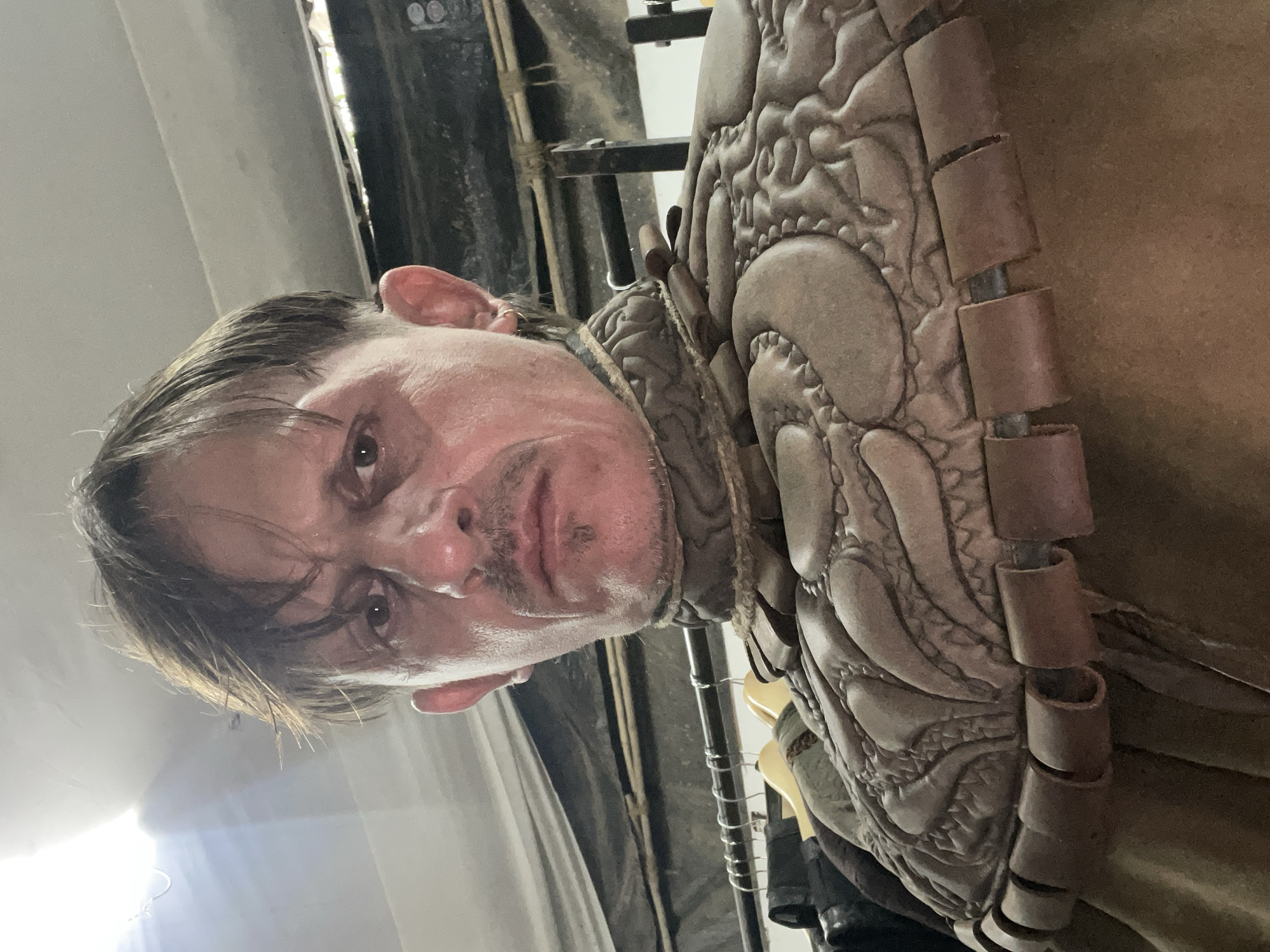The image shows a man wearing a textured, layered, leather-like costume. The man has fair skin, light brown hair that is slicked back, and appears to be perspiring. The costume is detailed with raised, patterned designs and cylindrical segments along the edges. The background is indistinct, suggesting a backstage or storage area with clothing racks and indeterminate structures. There is no indication of the location being Mumbai, India.