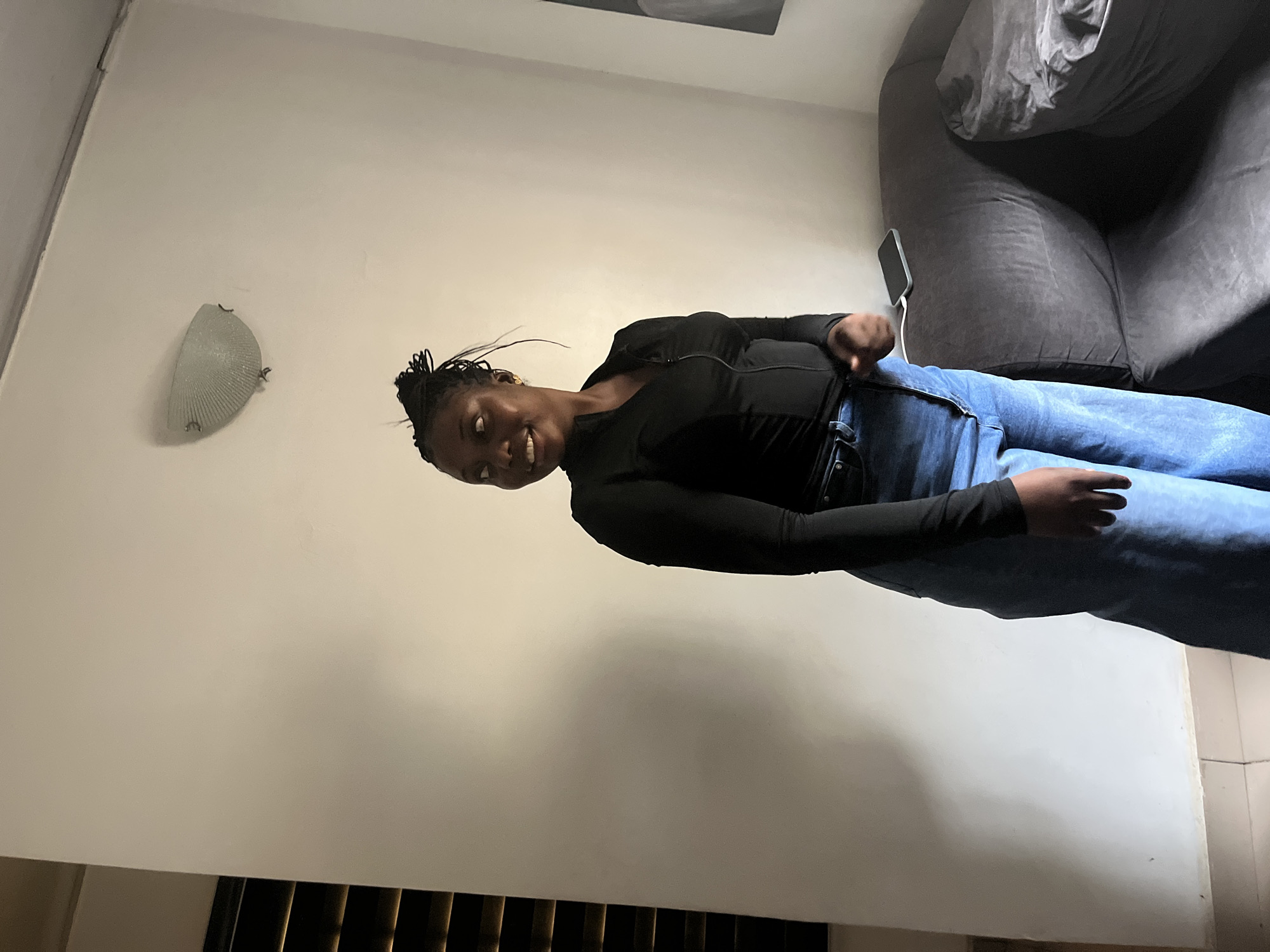 A young Black woman with dark braided hair styled in a bun stands in a sparsely furnished room. She is wearing a black, long-sleeved, form-fitting top with a zippered neckline and blue jeans. She is looking over her left shoulder and smiling. The room has plain white walls with a single light fixture mounted near the ceiling. To the left of the woman, there are blinds that suggest a window. To her right, a dark gray sofa is visible, with a smartphone resting on its surface. The overall impression is of a casual indoor setting. The scene is likely located in Abuja, Nigeria, given the context of the request.