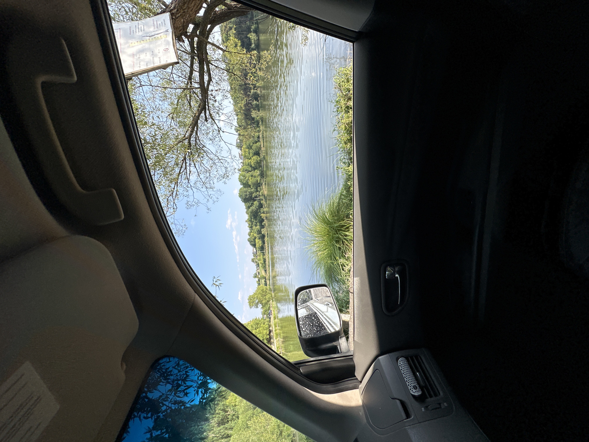 The image is taken from inside a vehicle, looking out the passenger window at a lake. The interior of the car is visible, including the door panel, side mirror, and a portion of the dashboard. The exterior shows a calm body of water with ripples, surrounded by lush green trees and vegetation. The sky is clear and blue with a few scattered clouds. A sign is attached to a tree on the right side of the frame, but its contents are not legible. There are no people or animals visible in the image. The overall scene suggests a peaceful, natural setting. The specific location in Makiv, Ukraine is not explicitly shown, but the natural landscape is consistent with a rural or park-like environment.