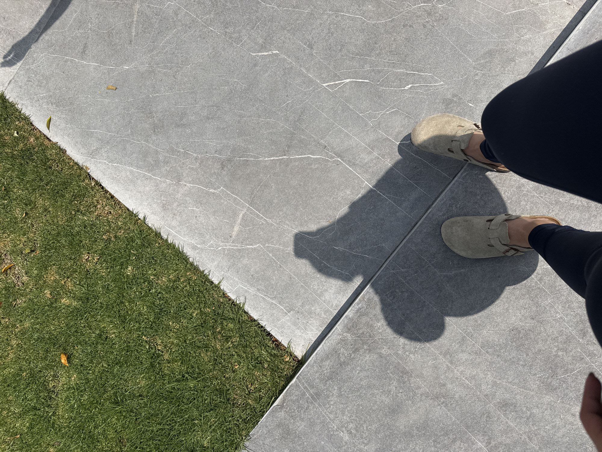 A high-angle, outdoor shot shows feet clad in tan Birkenstock sandals on gray, marble-patterned paving stones. The feet belong to a person wearing dark leggings. Adjacent to the paving stones is a strip of green grass. The shadows cast by the legs and feet are prominent in the image. No information from the image confirms the location in the city of San Bernardino Tlaxcalancingo, Mexico.