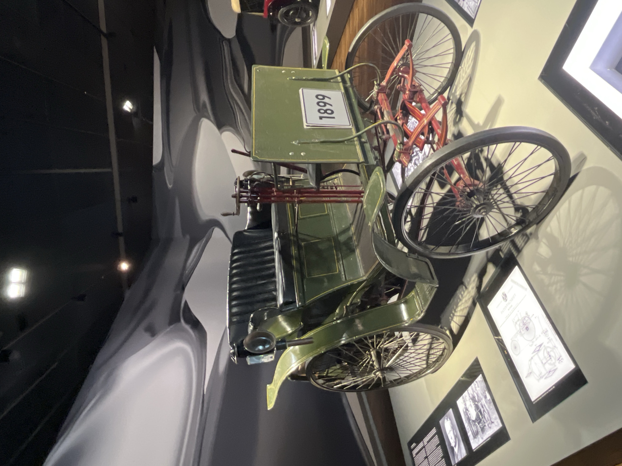 An antique green automobile, identified by a sign as from 1899, is displayed indoors, likely in a museum setting. The car has a bench seat upholstered in black, spoked wheels, and a visible steering column. It is positioned on a light-colored floor with several display panels showcasing historical images and diagrams. The background features a dark ceiling with lighting fixtures and a large, abstract mural with swirling grey and white patterns. The automobile is the central focus of the image, with other vehicles and display elements visible in the periphery. The scene suggests a historical exhibition, possibly within the Autostadt in Wolfsburg, Germany, given the prominent display of early automobiles.