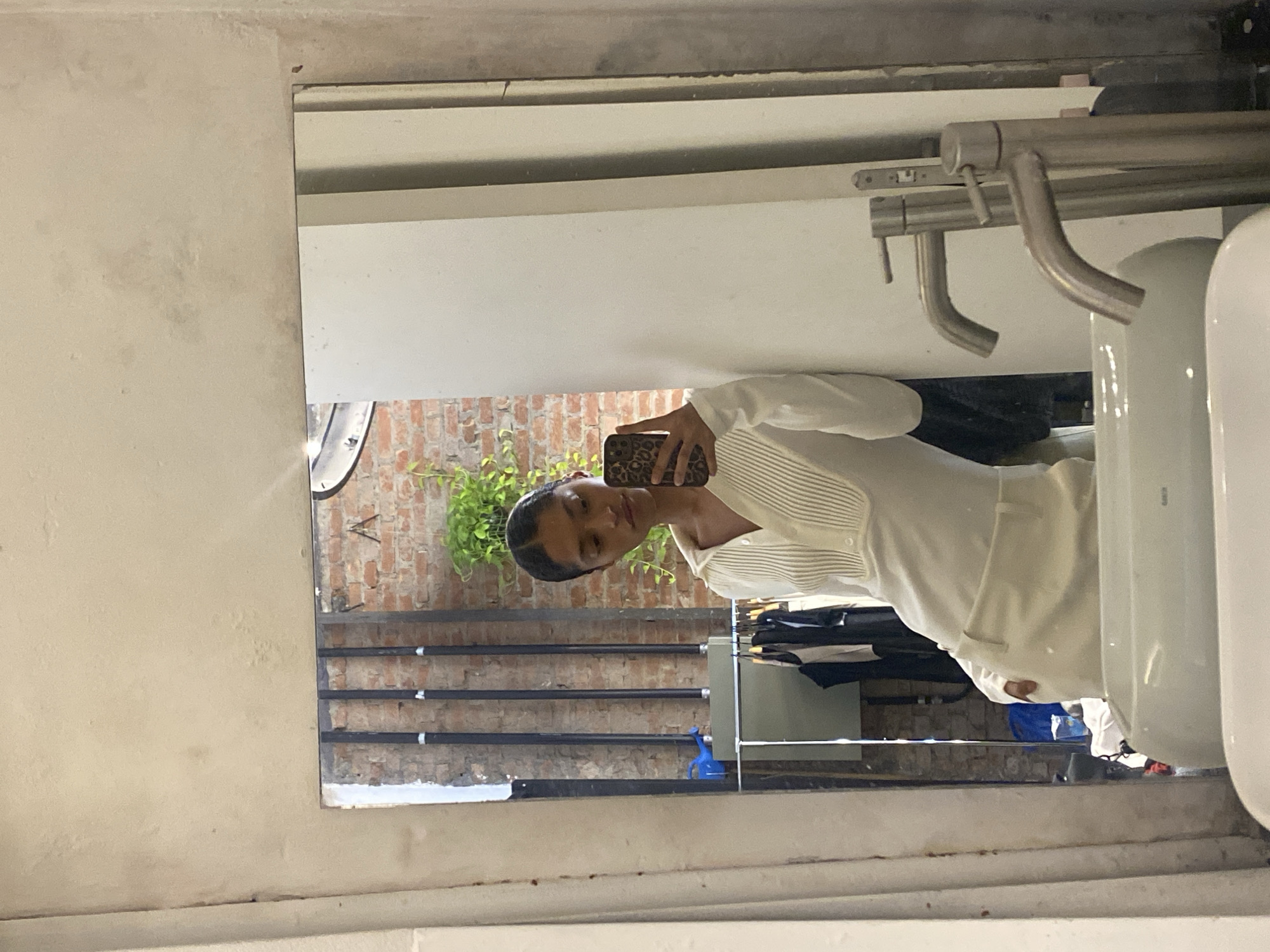 A person in a white outfit is taking a mirror selfie in what appears to be a commercial space, possibly a clothing store in São Paulo, Brazil. The reflection shows a brick wall with horizontal pipes, a green plant, and clothing racks with garments. The sink and plumbing fixtures in the foreground suggest the mirror is located in a restroom. The person is holding a phone with a leopard print case. The lighting is bright, and the overall scene is somewhat cluttered.
