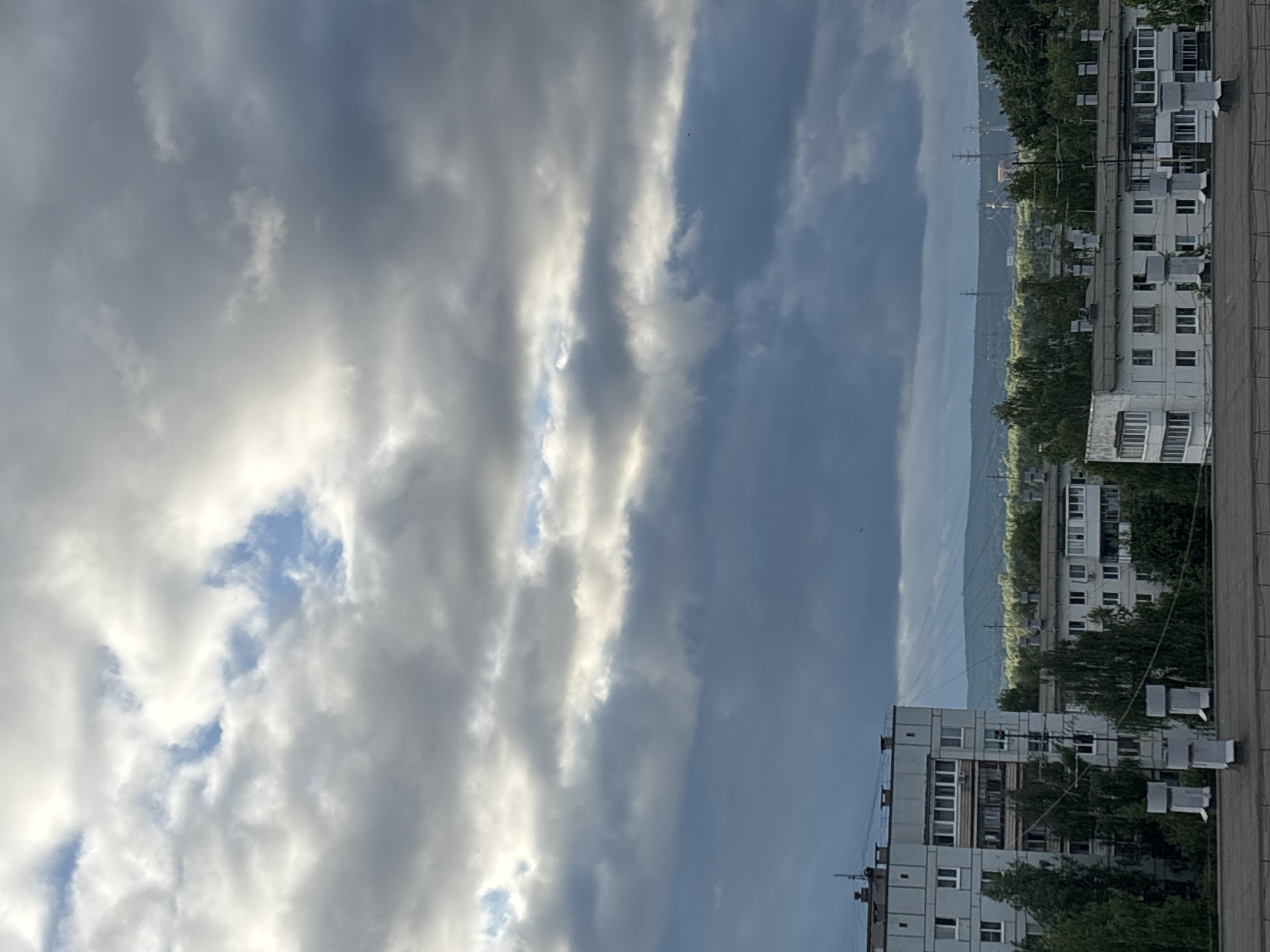 An outdoor photograph captures a residential urban landscape under an overcast sky in Chaikovsky, Russia. The scene is viewed from an elevated perspective, likely a rooftop, the edge of which is visible at the bottom of the frame with three light gray, rectangular ventilation or utility units.
The upper two-thirds of the image are dominated by a dense, multi-layered cloud cover, exhibiting shades of dark gray, bluish-gray, and bright white where sunlight illuminates the clouds. Patches of clear blue sky are visible in the upper left and upper right. Distinct crepuscular rays (sunbeams) descend from the clouds in the mid-left portion of the sky, extending towards the distant horizon.
In the midground, a prominent multi-story (approximately 5-7 stories) residential building, consistent with Soviet-era concrete panel architecture, occupies the left side of the frame. This light gray building features numerous windows and dark-framed balconies, one of which has a satellite dish. Multiple power lines or communication cables are visible traversing the space directly in front of this structure. To its right and further back, several other similar, light-colored residential buildings are visible, interspersed with mature deciduous trees displaying green foliage.
The background reveals a distant, undulating line of tree-covered hills or forests, appearing somewhat hazy against the sky. Above these hills, several tall, metallic electrical transmission towers (pylons) are discernible on the horizon. No human figures or active vehicles are visible within the frame. The overall lighting is subdued due to the extensive cloud cover, with intermittent brightness from the visible sunlight.