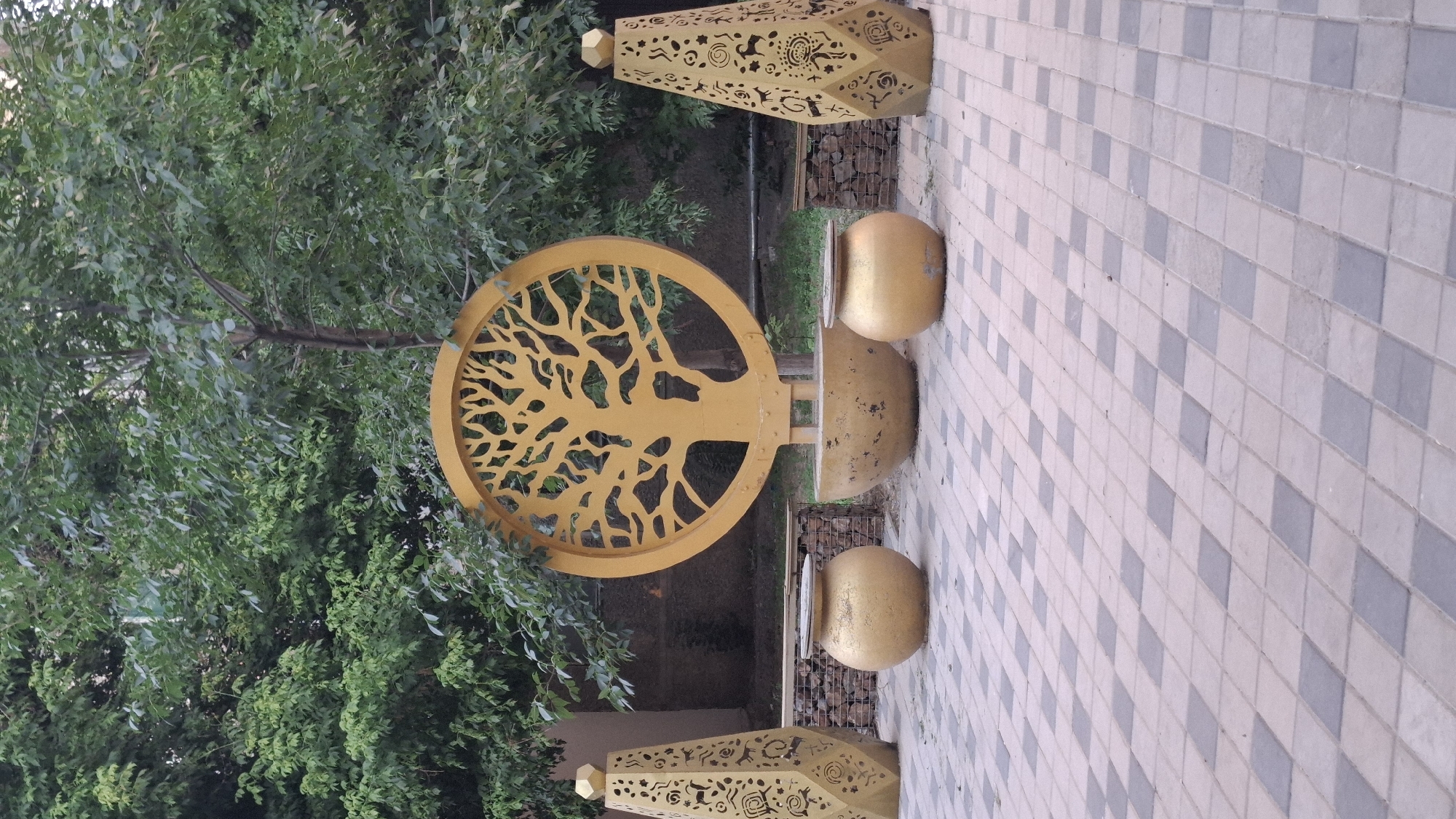 The image depicts an outdoor decorative installation in the city of Shymkent, Kazakhstan. In the center is a large, circular, golden metal cutout of a tree with intricate branches, supported by a stand. In front of the tree cutout is a large, shallow, golden, bowl-like planter. Flanking this central element are two tall, tapered, golden metal decorative pillars with cut-out patterns, and two spherical, golden planters. The installation is situated on a paved area composed of light and dark grey square tiles laid in a diamond pattern. Behind the installation, there is a low wall constructed from stacked stones, and further back, lush green foliage and trees are visible. The overall scene suggests a public space or courtyard designed for aesthetic appeal. There are no people or animals present in the image.