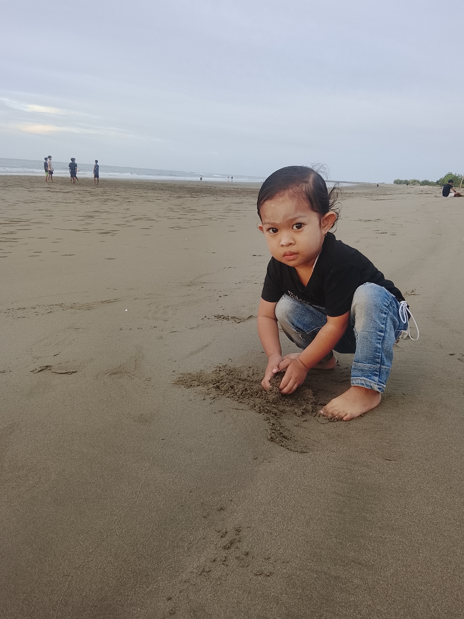 In Reudeuep, Indonesia, a young child is crouched on a sandy beach, playing with sand. The child, wearing a black t-shirt and blue jeans, is focused on the pile of sand in front of them. The beach extends to the ocean, where waves gently lap the shore under a cloudy sky. In the background, several people are visible walking and standing by the water, enjoying the coastal scenery. The overall atmosphere is one of a peaceful day at the beach.