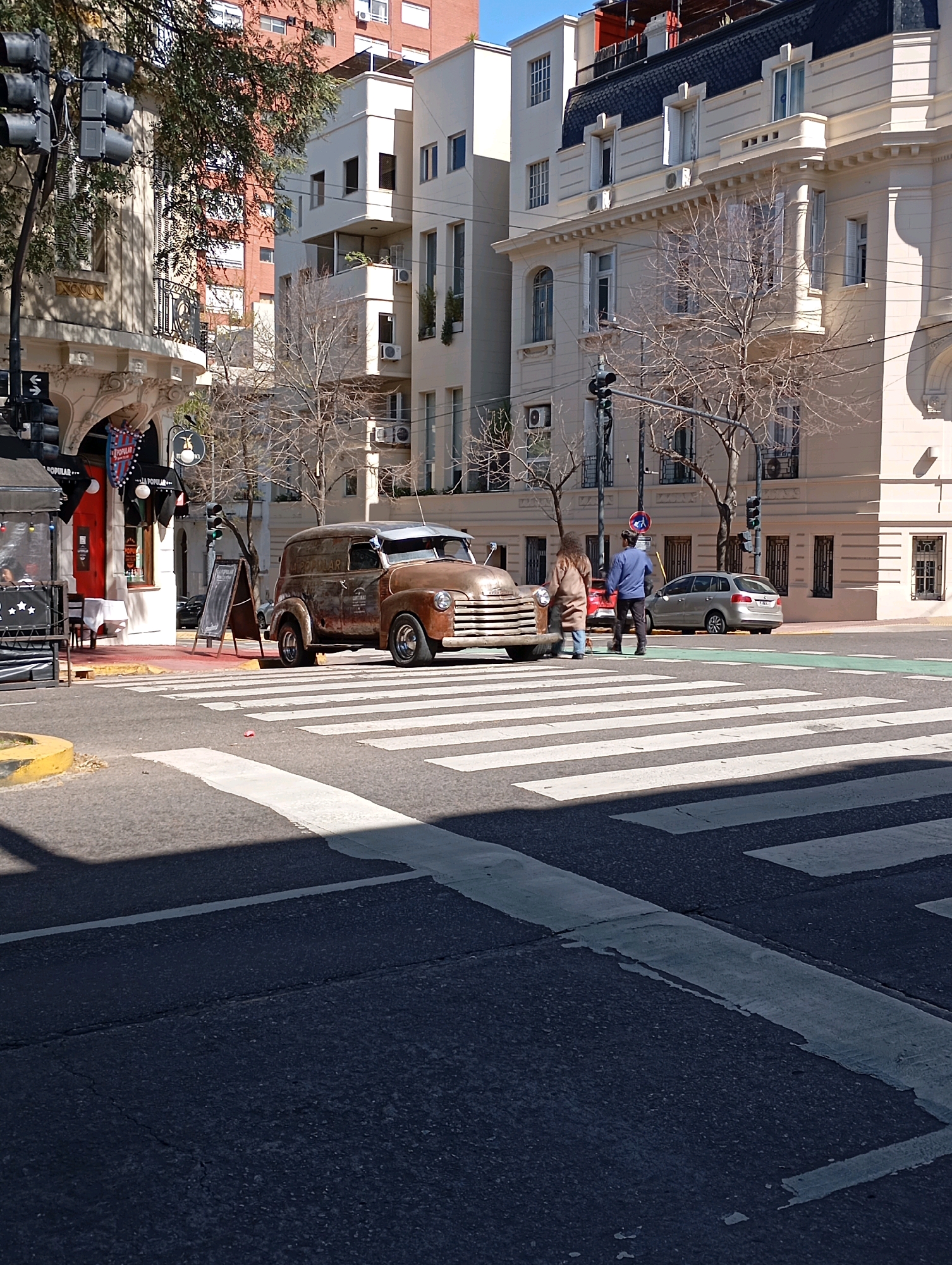 The image captures a street scene in the city of Buenos Aires, Argentina. In the foreground, a pedestrian crosswalk dominates the asphalt, marked with white stripes. To the left of the crosswalk, a yellow painted curb is visible. The main subject in the middle ground is a vintage, rusted brown panel van parked on the street, partially obscuring a portion of the crosswalk. Two individuals, one wearing a beige coat and the other a blue jacket, are standing near the front of the van, facing away from the camera and appearing to be engaged in conversation or observation.
Behind the van, a silver sedan is parked on the right side of the street. The background reveals a city environment with multi-story buildings. The architecture varies, with some buildings featuring lighter facades and balconies, while others display darker, more ornate roofs and window details. Trees with bare branches are scattered throughout the scene, suggesting a cooler season. Traffic lights and street lamps are also present, indicating an urban intersection. A small outdoor seating area with tables and chairs, likely part of a cafe or restaurant, is partially visible to the left of the frame.