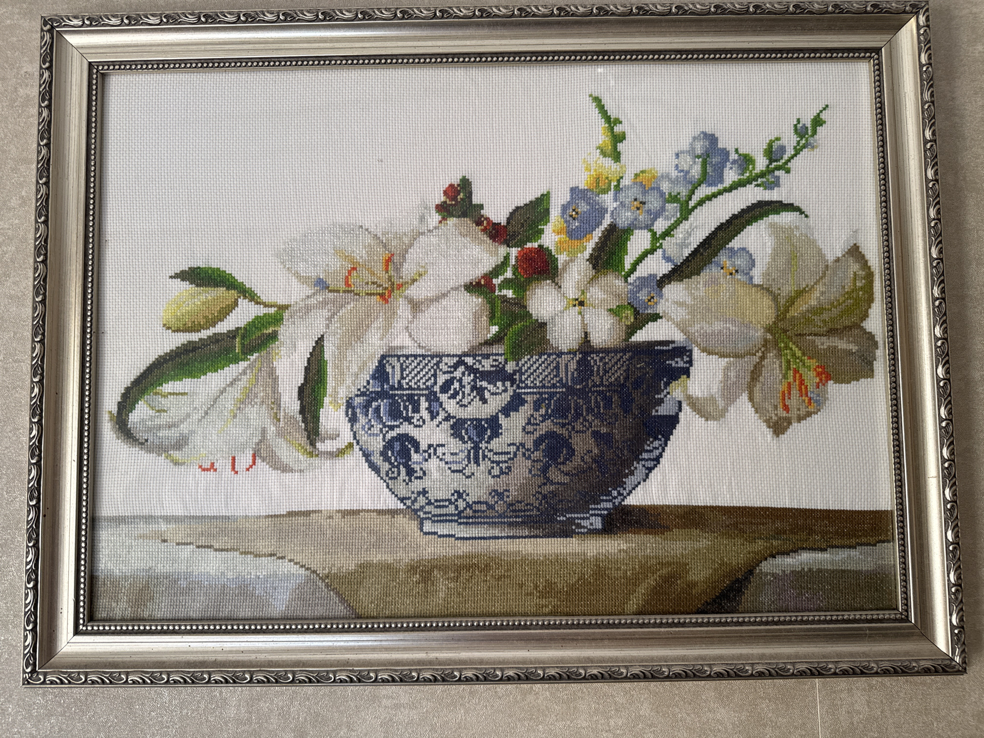 The image shows a framed cross-stitch artwork of a floral still life. The artwork depicts a bouquet of white lilies, smaller white flowers, blue flowers, and some red berries in a blue and white patterned vase. The vase is placed on a surface that appears to be a tablecloth or a draped fabric, which rests on a wooden surface. The background of the artwork is plain white. The frame surrounding the artwork is silver with decorative detailing. The artwork is displayed indoors, likely on a wall, as indicated by the lighting and shadow. There is no identifiable location in Bishkek City, Kyrgyzstan, as the scene is an indoor still life artwork.