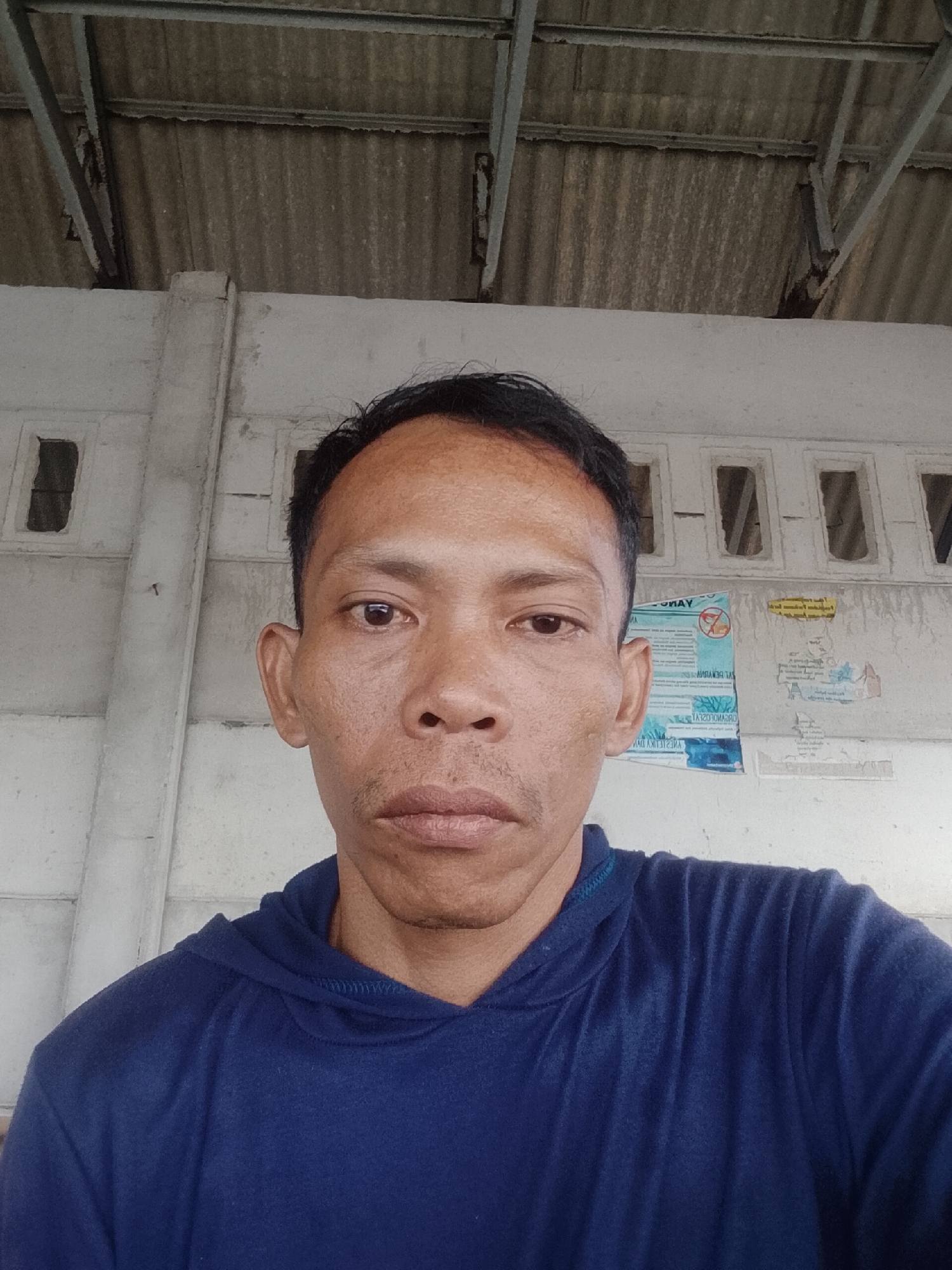 A medium, eye-level close-up captures a man in Bukit Ketok, Indonesia, dressed in a navy blue hooded sweatshirt. The man's face is centered in the frame, revealing medium brown skin, dark hair, and a neutral expression. Behind him, a concrete wall with repetitive, rectangular window openings is visible. The wall shows signs of wear, with posters and water stains adding texture. Above the wall is a corrugated metal ceiling. The scene lacks strong shadows, suggesting diffuse lighting.