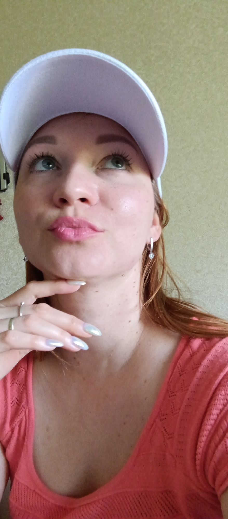 A woman wearing a white baseball cap and a salmon-colored top is looking up, her chin resting on her hand. She has red hair and pale skin with freckles. She is wearing earrings and rings. The background is a plain, light-colored wall. The location is Unknown, Unknown.