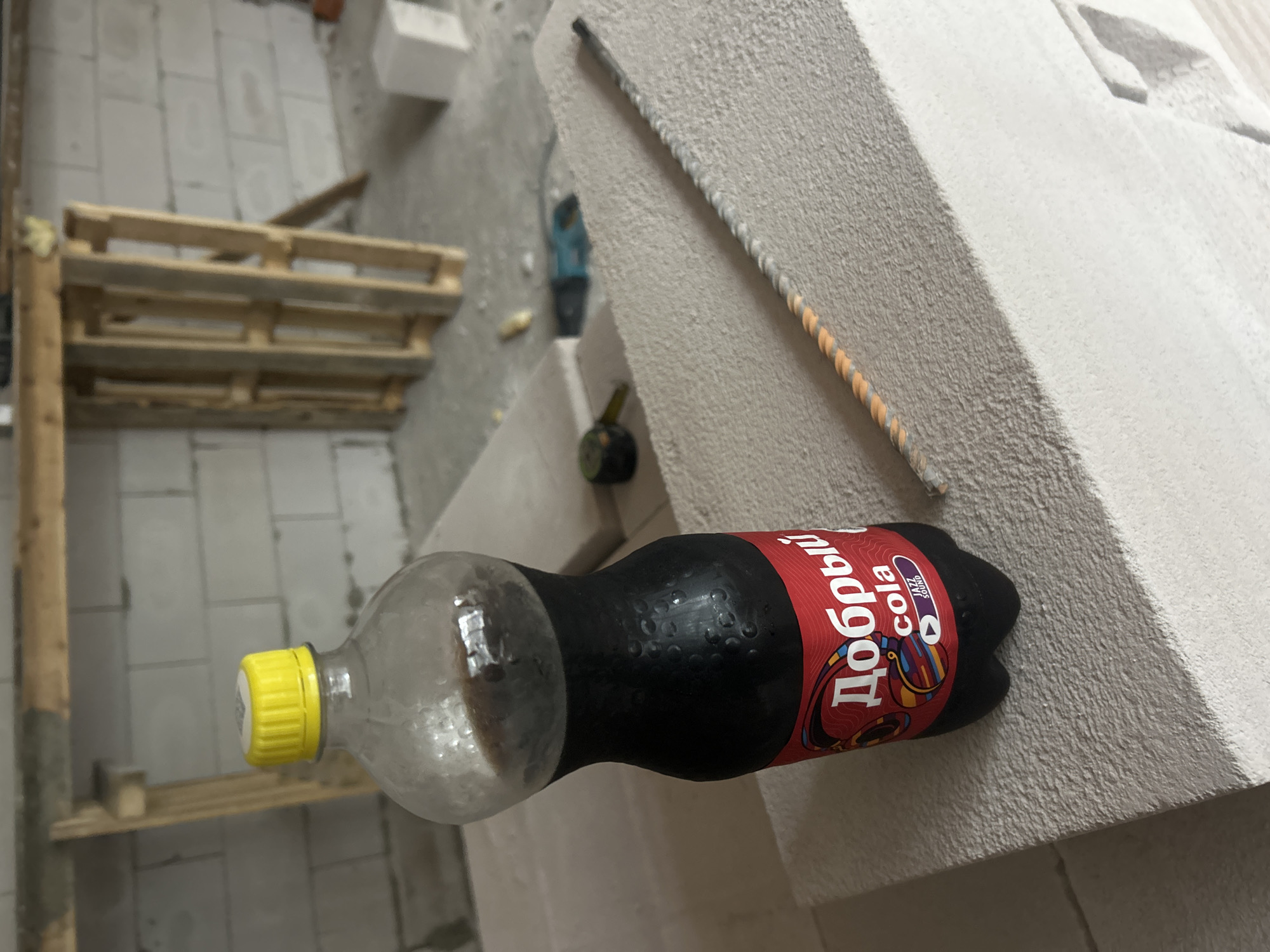 The image depicts a construction site in Saint Petersburg, Russia, characterized by unfinished cinder block walls, wooden scaffolding, and scattered building materials. In the foreground, a bottle of "Добрый cola" with a yellow cap rests on a stack of lightweight concrete blocks. Adjacent to the bottle is a piece of rebar. A tape measure and a power tool are visible in the background. The scene appears to be an indoor construction area with natural light entering from an unseen source.
