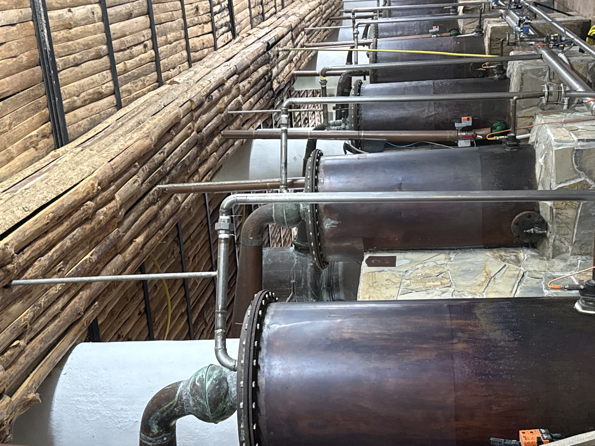 This image depicts an interior view of an industrial processing facility, likely a distillery, located in Tarnicheni, Bulgaria. The scene is dominated by a long row of large, cylindrical vats or stills, constructed from dark, oxidized copper or bronze, exhibiting varied patination and staining. The vats are supported by rough-hewn stone block bases of irregular shapes and colors.
A network of piping connects the vats. The foremost vat on the left features a curved copper pipe with significant green verdigris. This transitions to polished stainless steel piping, visible with bolted connections and flanges, extending along the row. Vertical and horizontal stainless steel pipes further connect various components. Numerous valves, gauges, and electrical components, including orange-cased units, are attached to the lower sections of the vats and associated plumbing. A bright yellow hose is partially visible on the floor in the background.
The ceiling is constructed from numerous rough-hewn wooden logs or branches, laid horizontally and supported by darker, possibly metal, beams. A white-painted wall is visible on the left, while a textured stone wall with a dark brown rectangular sign bearing the number "2" (or "Z") is visible between two vats on the right. No individuals are present in the scene.