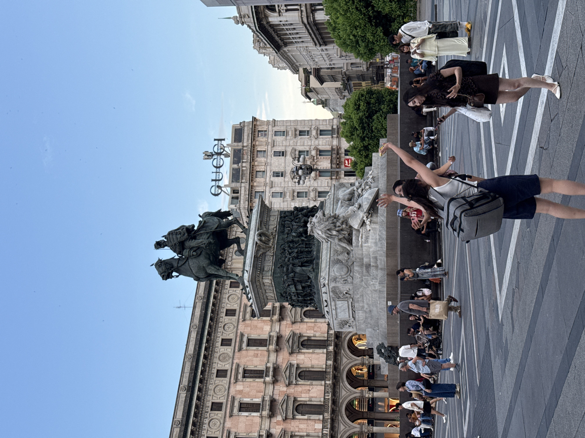 The image depicts a wide-angle shot of a public square in Milan, Italy, likely Piazza del Duomo, given the presence of significant architecture and a prominent equestrian statue. In the foreground, several people are captured in casual poses. Two women in the immediate foreground appear to be interacting, one with her arms raised. Behind them, a diverse group of individuals are scattered throughout the plaza, some walking, others seated.
Dominating the mid-ground is a large, elaborate monument featuring an equestrian statue of a man in military attire, mounted on a horse. The base of the monument is richly adorned with sculpted reliefs. Behind the monument, several grand buildings with classical architectural elements are visible. One building prominently displays a large "GUCCI" logo. Green trees provide a touch of nature amidst the urban landscape. The sky above is a clear, pale blue, suggesting it is late afternoon or early evening. The paving of the square is made of large stone slabs with white lines demarcating sections. The overall atmosphere appears to be one of a busy, yet relaxed, urban gathering space.
