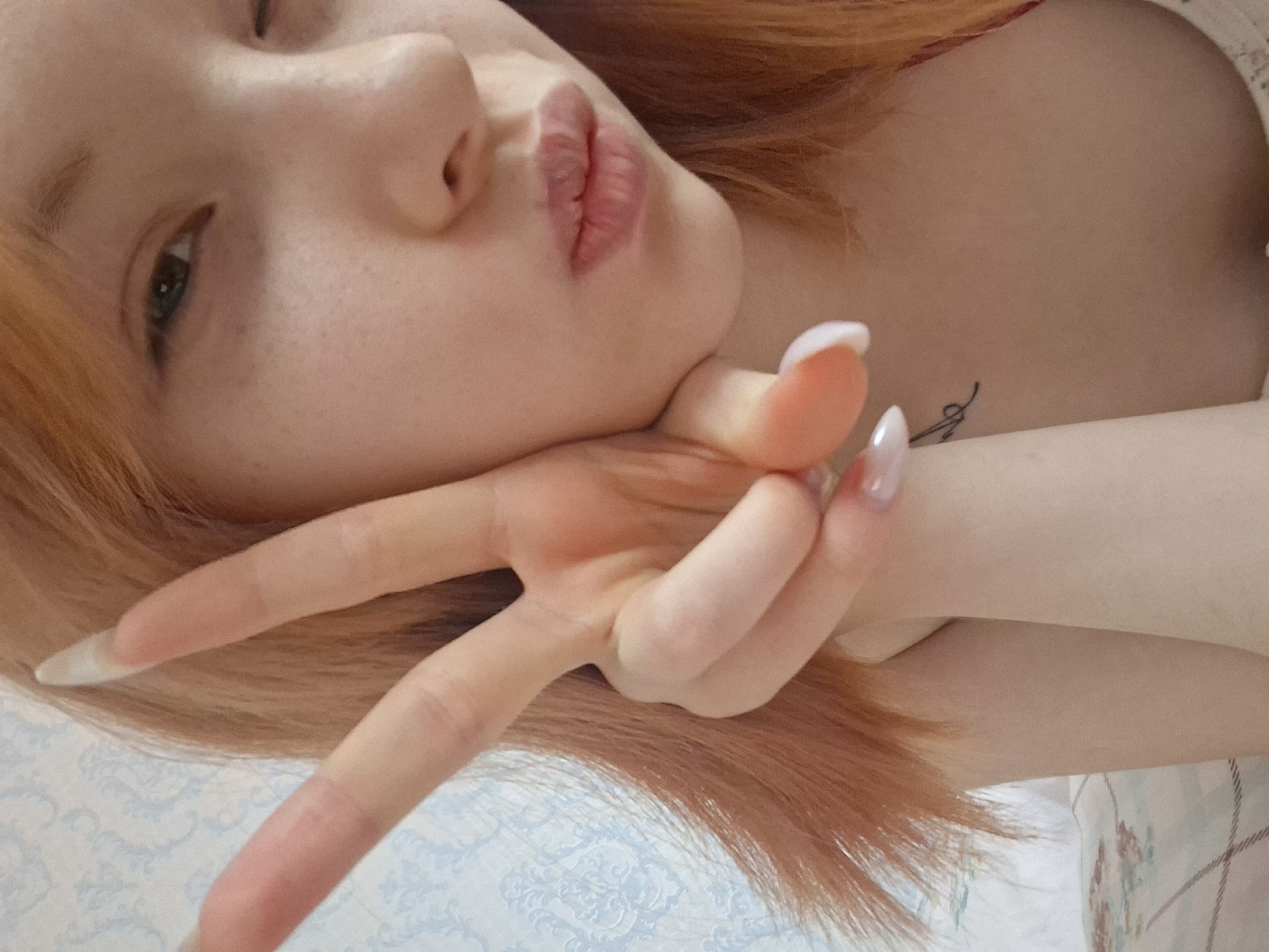 A close-up, angled photograph features a young woman with light skin and freckles. She has orange-red hair and is making a kissing face. Her left eye is visible and brown. She is holding her hand up in a peace sign, her fingers extended, with long, manicured nails. A small tattoo is visible on her chest. The background is blurry, with patterned fabric. The location in the city of Chaikovsky, Russia is not verifiable from the image.