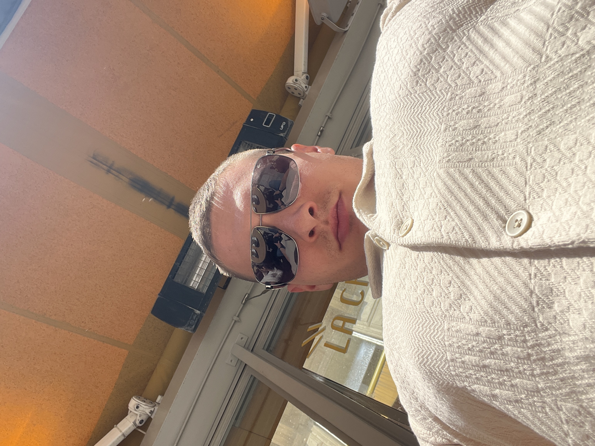 A man in sunglasses is captured in a close-up, upward-angled selfie, likely taken on a bright, sunny day. He is seen from the chest up, wearing a light cream or beige collared shirt with a distinct textured, possibly patterned, weave and visible buttons down the placket. His face is partially obscured by large, mirrored aviator-style sunglasses, which reflect an interior setting.
The man's head is tilted slightly. The background indicates he is in an indoor or covered outdoor space, possibly a cafe or restaurant in Paris, France. Above and behind him, a ceiling made of light brown, possibly cork or textured acoustic panels, is visible. Several fixtures are mounted overhead, including what appears to be a black rectangular patio heater or speaker with "UFO" faintly visible on its side, and white metal structural supports.
Through a large glass window or door behind the man, golden text spelling out "LA CA" is clearly visible, likely part of a business name such as "LA CASA" or "LA CAFE." The reflections on his sunglasses show fragmented glimpses of an indoor scene, possibly containing other people or interior elements. The overall lighting is bright, with strong sunlight creating highlights on his shirt and the surrounding structures, suggesting a clear sky. The man's expression, though largely hidden by the sunglasses, appears neutral.