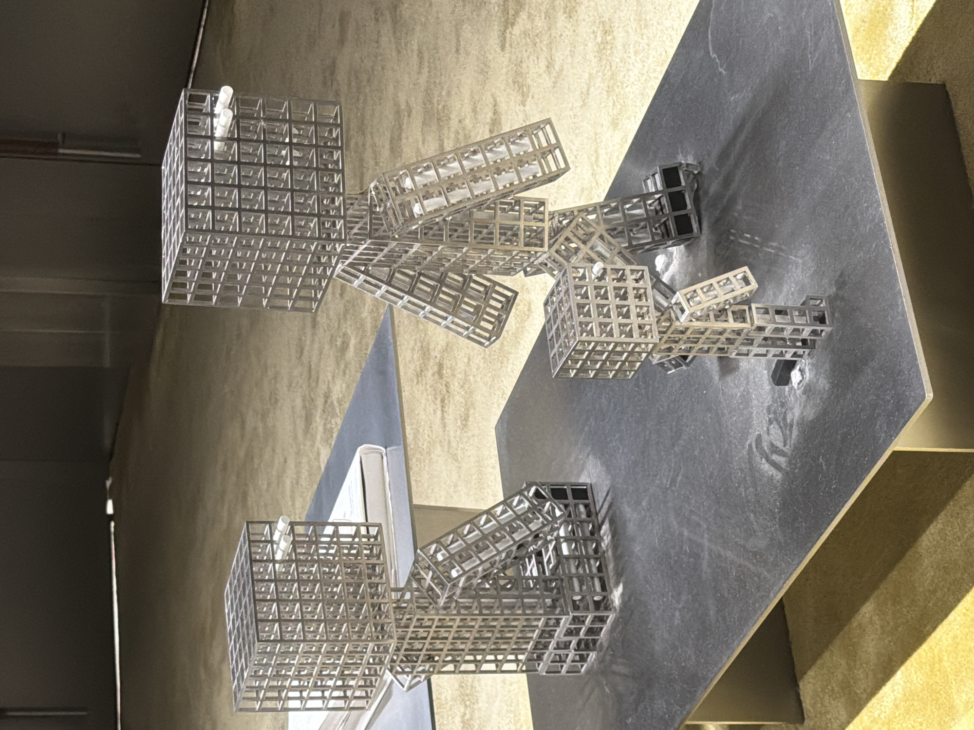 Three abstract sculptures resembling Minecraft characters are displayed on a dark gray, rectangular table. The sculptures are constructed from a grid-like material, appearing to be metal. They are positioned in a staggered arrangement, with the largest sculpture in the background, a medium-sized one to its left, and the smallest in the foreground. The environment suggests an indoor setting, possibly a gallery or showroom, with a light-colored carpeted floor visible in the background. The lighting creates shadows that extend from the sculptures across the table. The sculptures do not exhibit any discernible interactions.