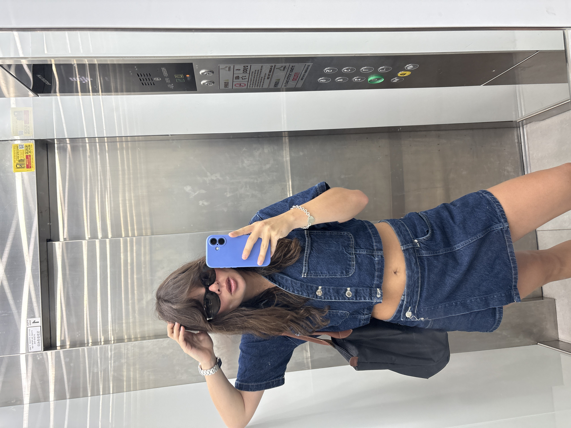 A woman is taking a mirror selfie in an elevator. She has medium-length brown hair, is wearing dark sunglasses, and is dressed in a denim crop top and a denim skirt. She is holding a purple smartphone in her right hand. The elevator has a brushed metal interior and a panel of buttons on the right side, some of which are illuminated. The elevator is located in Tel-Aviv, Israel, as indicated by the Hebrew text on the elevator panel. The overall scene suggests a casual, urban setting.