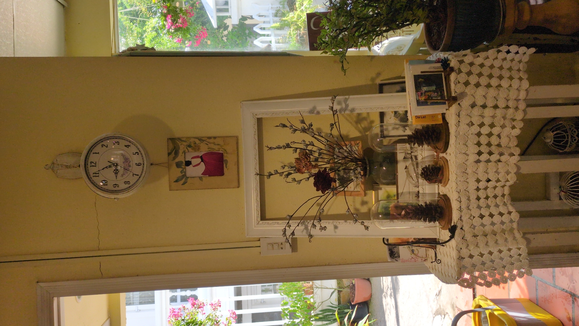 The image shows a room with a yellow wall. Hanging on the wall are a white clock and a framed picture. Next to the framed picture is a white frame with dried flowers inside. On a shelf next to the frame is a book titled "Da Lat" and three glass domes containing pine cones. A white lace tablecloth hangs over the shelf. Outside the window are flowers and a white picket fence. The location is likely Da Lat, Vietnam, given the book title.