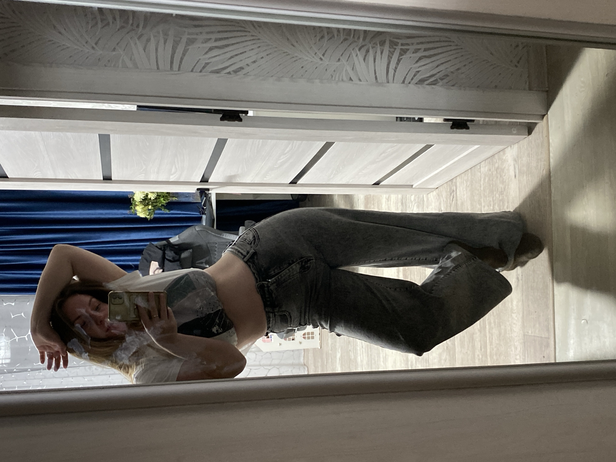A young woman is taking a selfie in a mirror. She is wearing a white t-shirt and gray jeans. The background shows a room with blue curtains and white furniture. The location is potentially Noviy, Russia.