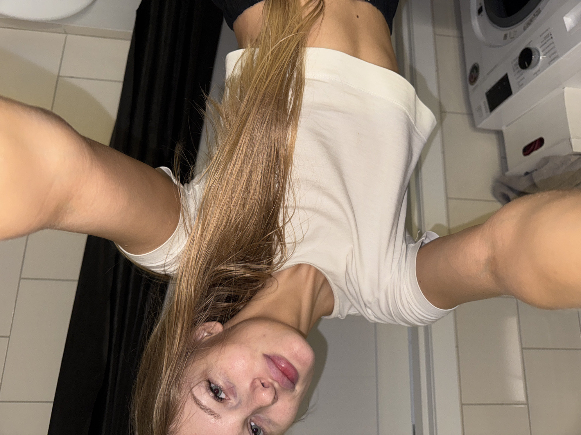 A young woman is taking a selfie in what appears to be a bathroom. She has long, light brown hair and is wearing a white crop top. Her face is turned towards the camera, and she has a neutral expression. In the background, a washing machine is visible, along with tiled walls. The overall setting suggests a domestic environment. There is no specific indication of the city of Perm, Russia.