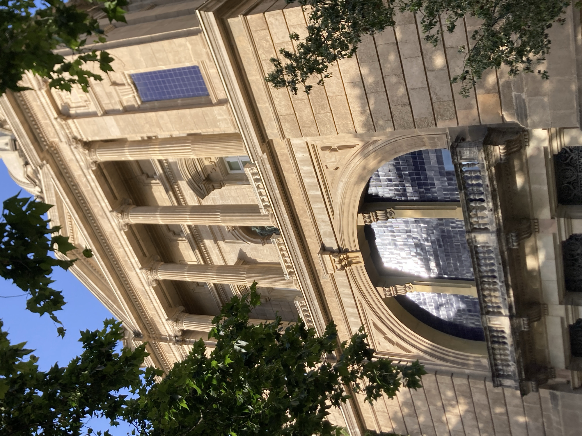 The image shows the facade of the Palau de la Generalitat de Catalunya, a historical building located in the Gothic Quarter of Barcelona, Spain. The building's exterior is characterized by its classical architecture, featuring a prominent portico with Corinthian columns, a balustrade, and arched windows. The stone facade is a warm, sandy color, illuminated by natural sunlight, creating shadows that accentuate the architectural details. Lush green foliage from trees in the foreground partially obscures the view, adding a natural element to the urban landscape. The sky above is a clear, bright blue. There are no people or discernible activity visible in the image.