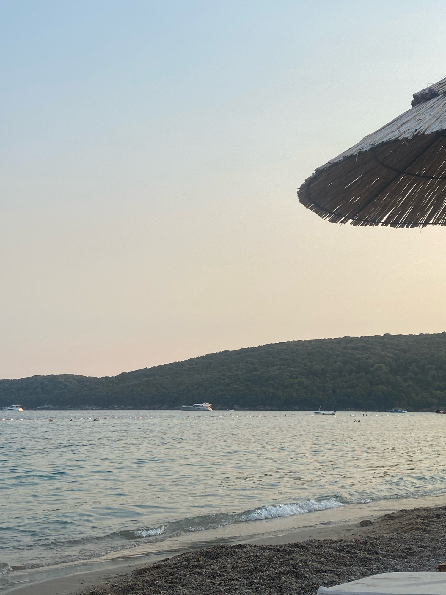 The image depicts a serene coastal scene, likely taken at a beach near Prijevor, Montenegro, during the late afternoon or early evening. In the foreground, a pebble and sand beach stretches towards the calm, rippling water of the sea. A portion of a thatched umbrella is visible in the upper right corner, casting a shadow that is not captured in the frame.
The middle ground is dominated by the tranquil sea, with a line of buoys marking a boundary in the distance. Several boats, including yachts and smaller vessels, are anchored or cruising on the water. Some individuals can be seen in the water, suggesting recreational activities.
The background features a lush, tree-covered hillside that slopes down to meet the sea. The sky above is clear with a soft, gradient of light blue and pale yellow, indicating either sunrise or sunset. The overall atmosphere is one of peacefulness and natural beauty.