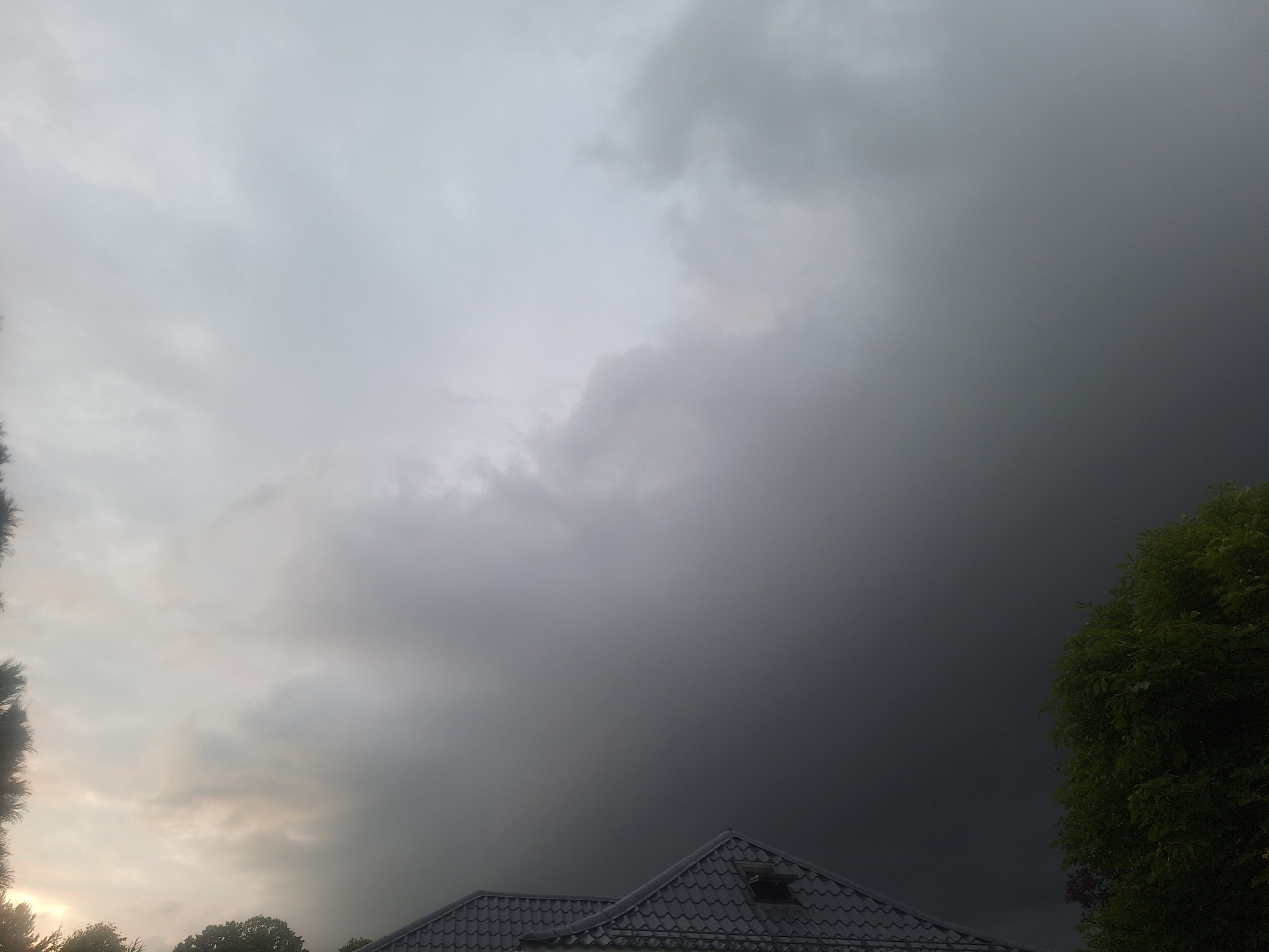 A wide shot captures a dramatic sky over a residential area in Mishutkino, Russia. The sky is dominated by heavy, dark clouds that suggest an impending storm, with lighter, overcast conditions visible towards the left. In the foreground, the dark, tiled roof of a house is visible, featuring a small skylight. To the right, the lush green foliage of a tree extends into the frame. The scene is devoid of people or any discernible activity, presenting a static, atmospheric view of the weather.