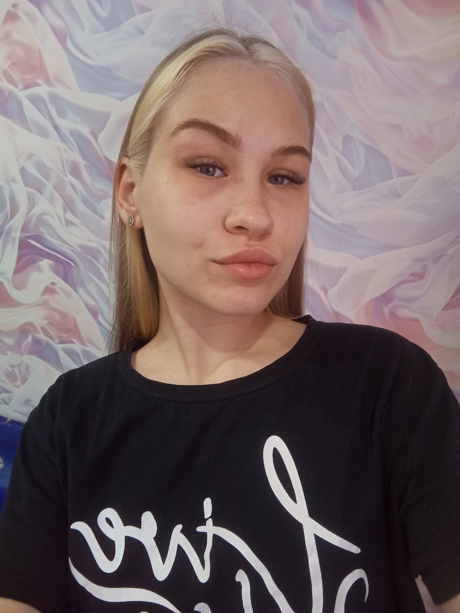 A young woman is depicted in an indoor portrait, looking directly at the camera with a neutral expression and a slight pout. She has long, straight, light blonde hair, appearing to have darker roots or streaks, and light blue or grey eyes. Her eyes are accentuated by prominent, dark eyelashes, and her eyebrows are well-defined. Her fair skin shows natural texture. She wears small, round, silver-toned stud earrings with a decorative, possibly crystalline, design.
She is dressed in a plain black, crew-neck t-shirt. On the front of the shirt, partially visible white stylized script lettering can be seen, including characters resembling "o", "u", and a decorative swirl, but the complete word or phrase is obscured.
The background features a soft, flowing, wavy pattern in pastel shades of purple, pink, and white, resembling a decorative wall covering or draped fabric. The lighting is even, illuminating the subject clearly. The image was taken in Krasnodar, Russia.