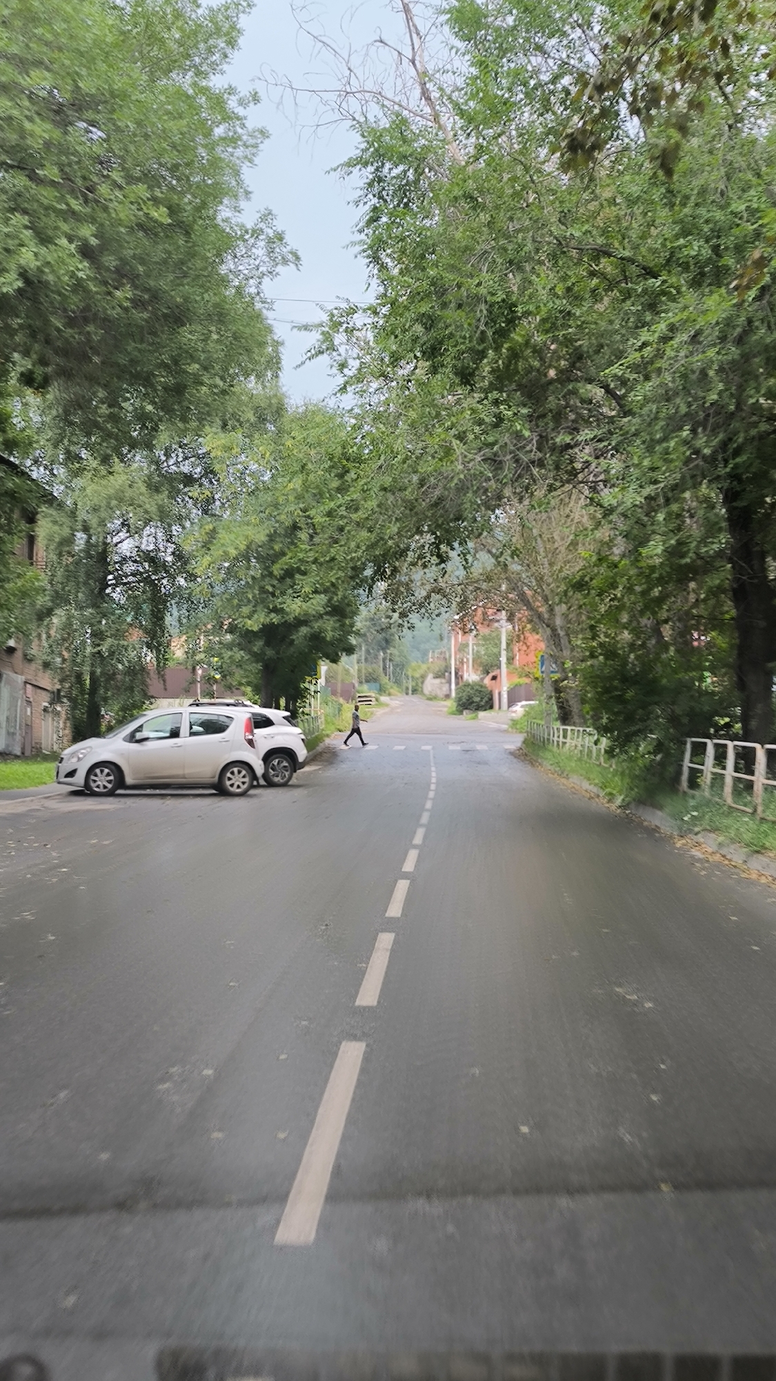 A person is walking across a street in Miass, Russia, on a cloudy day. The street is wet, suggesting it has recently rained. Two cars are parked on the left side of the road, and a pedestrian crossing is visible in the distance. Trees line both sides of the street, and a building is visible on the left. The scene appears to be a quiet residential area.