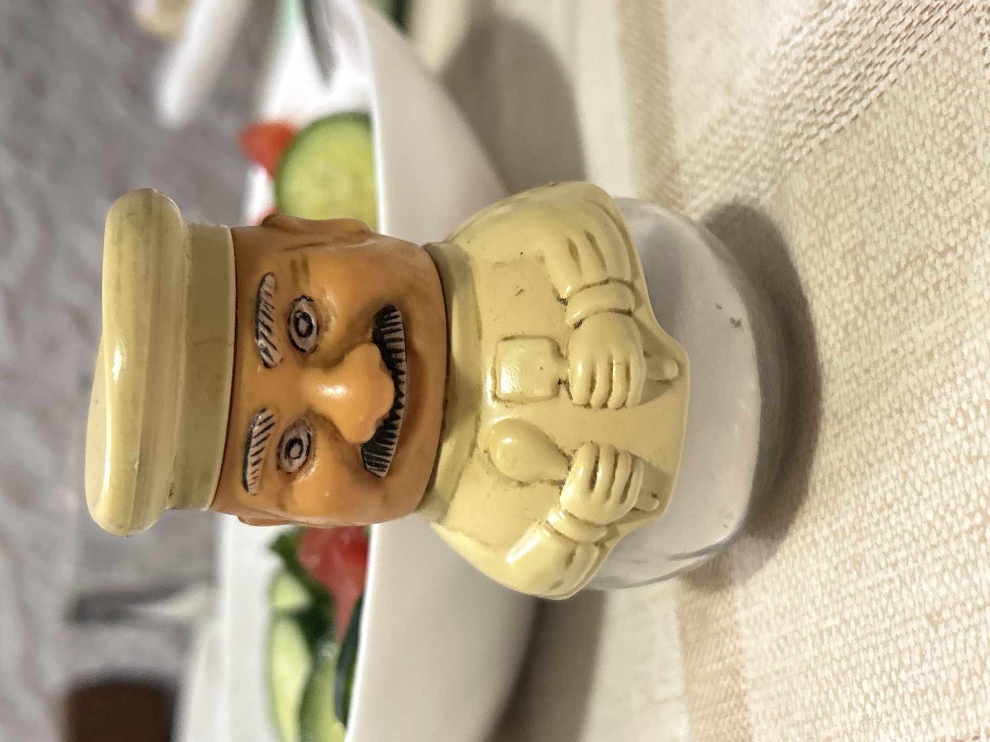 A close-up image depicts a chef-shaped salt shaker made of painted ceramic. The chef figure is light brown with a yellow toque and jacket, with simulated crossed arms holding a spoon and knife. The chef’s face has exaggerated features and black detailing. The figure is attached to a clear glass base. In the background, out of focus, is a plate of salad with tomato and cucumber. The image appears to be taken on a table covered with a beige textured tablecloth. The location in the city of Cherven, Bulgaria, is not apparent from the visual information provided.