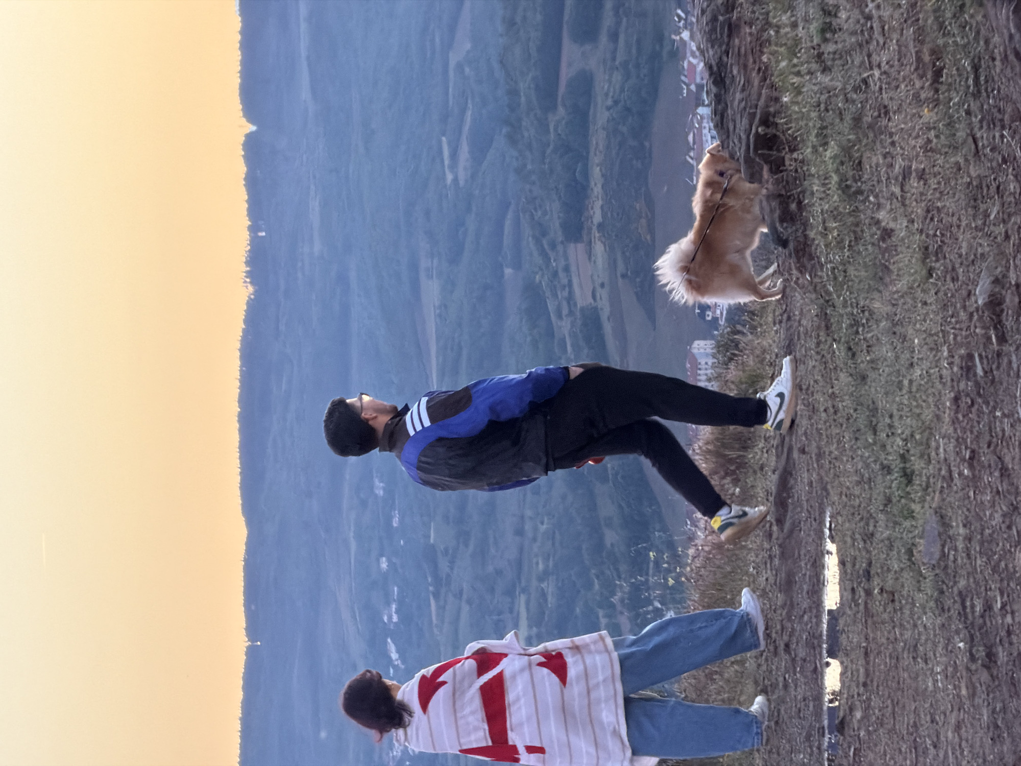 Two people and a dog are standing on the edge of a hill overlooking a valley in Bragança, Portugal. One person is wearing a blue and black jacket and black pants. The other person is wearing a white and red striped shirt and blue jeans. The dog is small and fluffy. The valley below is green and hilly, with buildings in the distance. The sky is a light orange color.