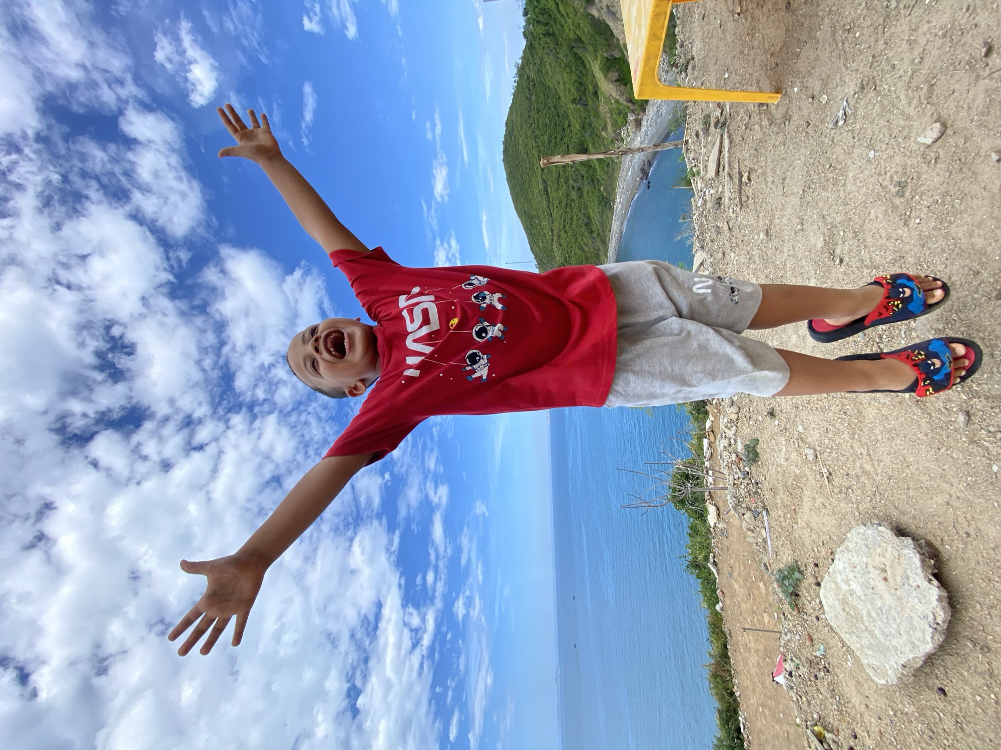 A young boy in a red NASA t-shirt and grey shorts jumps with his arms outstretched against a backdrop of a bright blue sky with scattered white clouds, the ocean, and a lush green hill. The boy's mouth is open in a wide, joyful expression. He is wearing black sandals with a red trim, adorned with cartoon characters. The setting appears to be a slightly rugged, elevated area overlooking a coastal bay, possibly in or near the city of Nha Trang, Vietnam, given the presence of the ocean, hills, and the general landscape. To the right, a bright yellow table is visible, suggesting a casual outdoor seating area. The ground is covered in dirt, pebbles, and some scattered debris. The ocean stretches out to the horizon, with a few small boats visible in the distance.