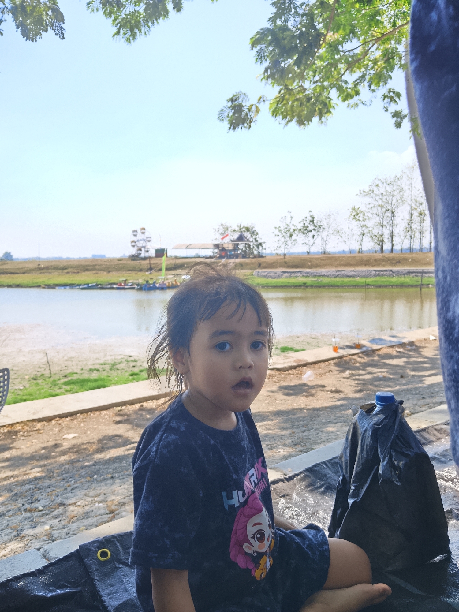A young Indonesian girl, with dark hair and wide eyes, sits on a black tarp near a body of water. She is wearing a dark t-shirt with a cartoon character and the word "HURK" on it. Beside her is a large black plastic bag with a blue bottle cap on top. The background shows a river or lake, a sandy shore, and trees. In the distance, a Ferris wheel and some buildings are visible. The scene appears to be a park or recreational area in the city of Ngadrin, Indonesia.