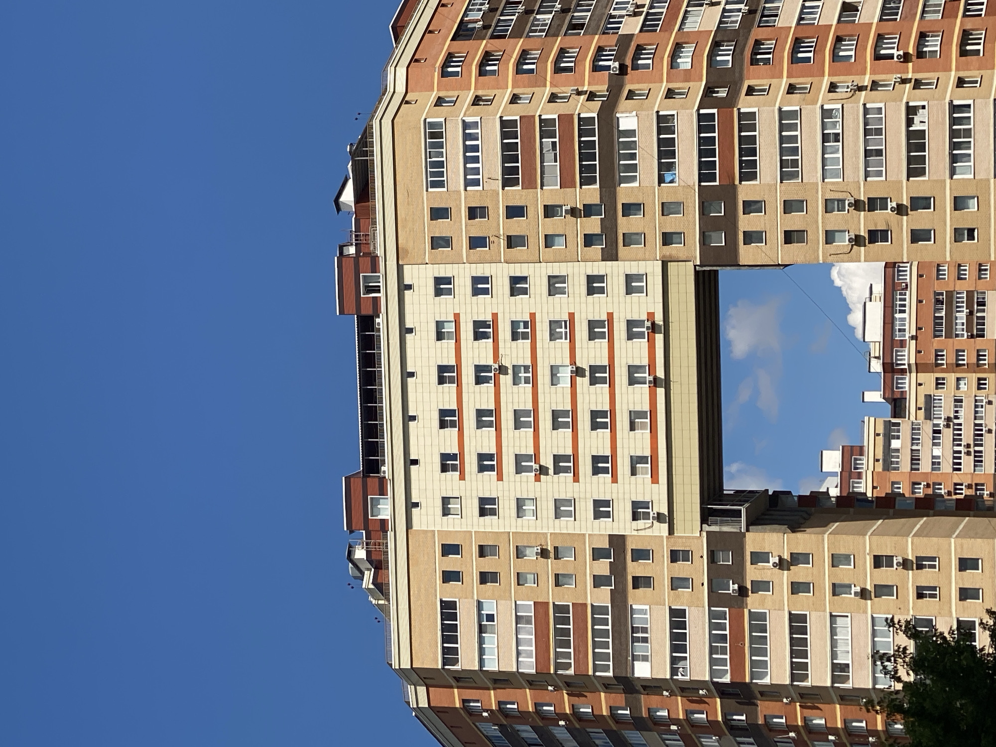 This media file showcases a large, multi-story building with a unique architectural design featuring a central open space. The building is predominantly light brown and beige with accents of reddish-brown and white. Numerous windows are visible across the facade, arranged in regular patterns. Some windows are rectangular, while others appear to be square. Several windows have air conditioning units installed outside.
The central open section of the building reveals a clear blue sky with a few white clouds, suggesting fair weather. The architecture is modern, with projecting balconies and tiered sections. The building is viewed from a low angle, emphasizing its height against the sky. The orientation of the image is such that the building appears to be sideways, and the sky occupies a significant portion of the frame.
The scene appears to be a residential or office building complex. The lighting suggests it is daytime, likely late morning or afternoon, given the bright sunlight and clear sky. There are no people or distinct objects in the foreground or on the balconies, focusing the attention solely on the building's structure. The overall impression is one of urban architecture. No visible text is present in the image. The location context is Kotelniki, Russia.