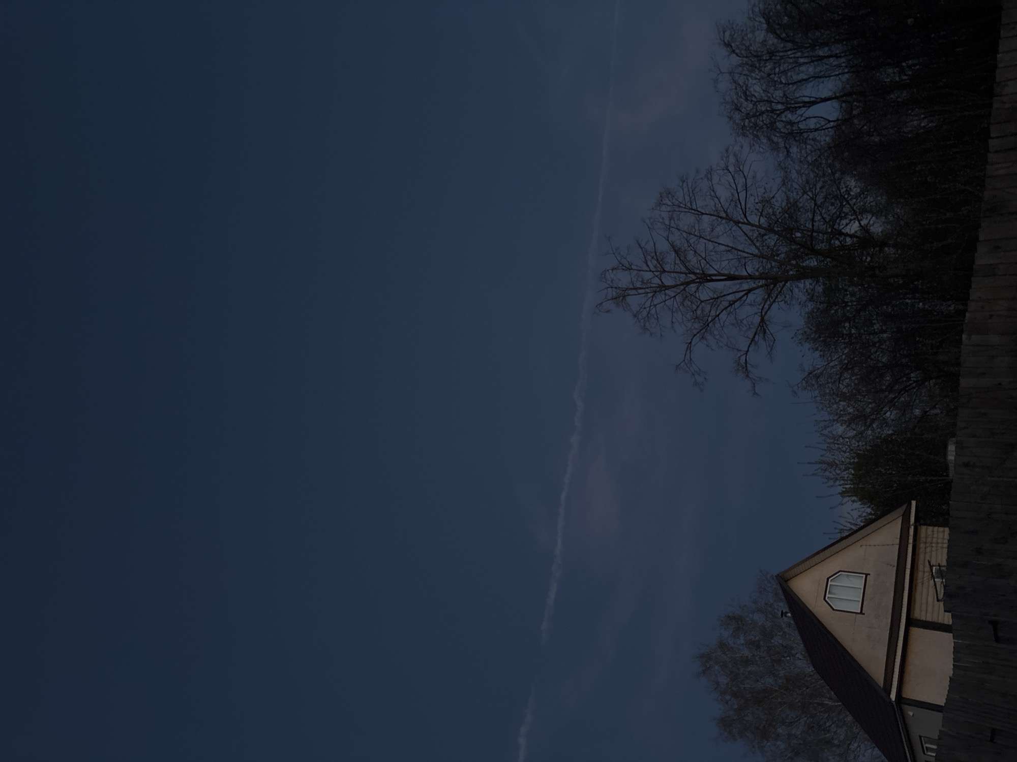The image shows a night view in Фоки, Russia, including a portion of a house with a triangular roof and an arched window on the upper floor, bare trees against a dark blue sky, and a contrail. The photograph is taken from a low angle, looking upwards. A fence is also visible.
