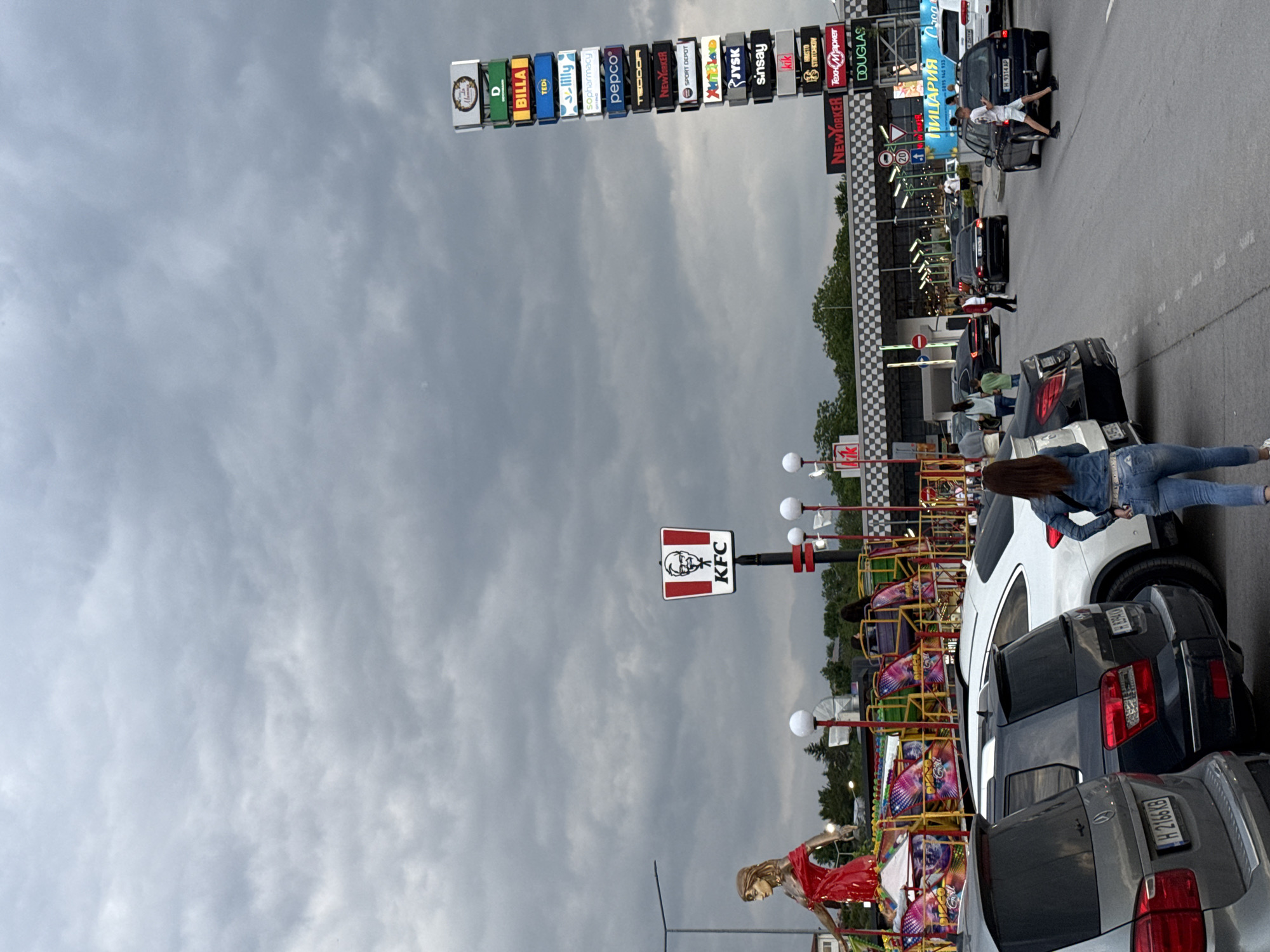 The image shows a street scene in Shumen, Bulgaria. Several cars are parked along the right side of the road. A woman in jeans and a denim jacket is standing next to a white SUV. A small children's carnival ride is visible behind the parked cars. Above the road is a sign displaying the logos of various businesses, including Billa, Tedi, Lilly, Sopharmacy, Pepco, Teodor, New Yorker, Sport Depot, JYSK, Sinsay, Kik, Hristo Stintchkov, Texn Market, and Douglas. Further down the road, another sign features a KFC logo. In the distance, there are more cars, buildings, and pedestrians. A sign that reads “Пицария Сса” with the numbers “5895 140 933” appears next to a black vehicle. The sky is overcast.