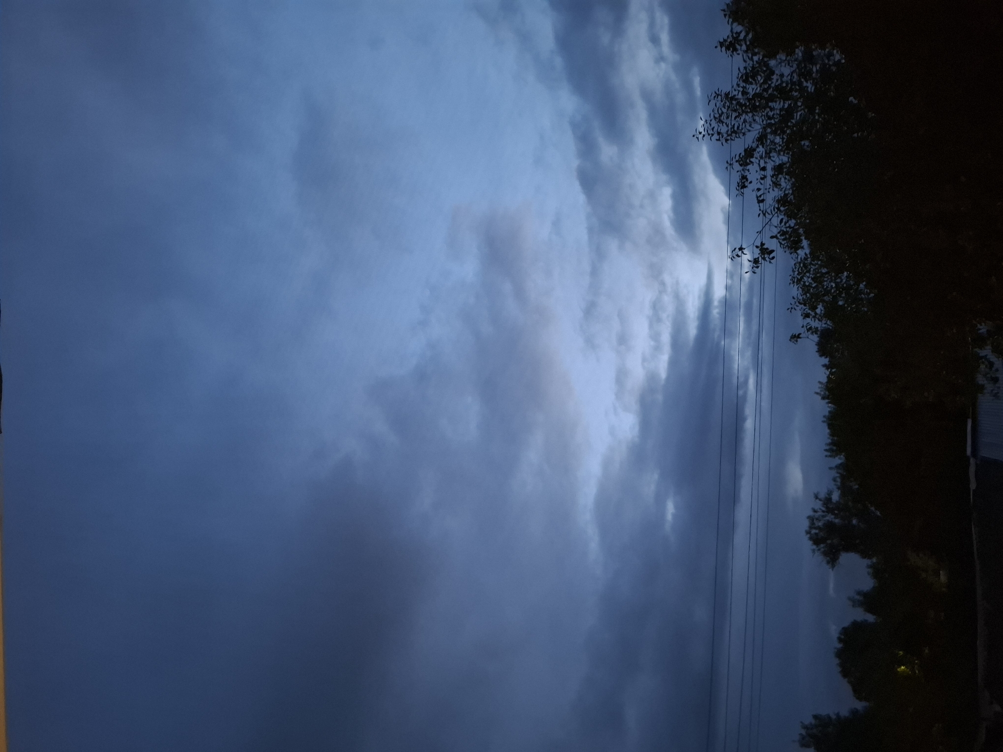 The image captures a dusky sky over a silhouetted landscape of trees and power lines. The sky is filled with a mix of dark and lighter gray clouds, suggesting either approaching or receding twilight. A hint of pale light is visible through a break in the clouds in the upper right quadrant. In the foreground, the silhouettes of dense trees and bushes obscure much of the ground, with some faint lights possibly from residential areas visible within the foliage on the left. Several horizontal power lines stretch across the middle ground, intersecting the view of the trees and sky. The overall impression is one of a serene, possibly humid, evening in a semi-rural or suburban area. The location is identified as Novlyanka, Russia.