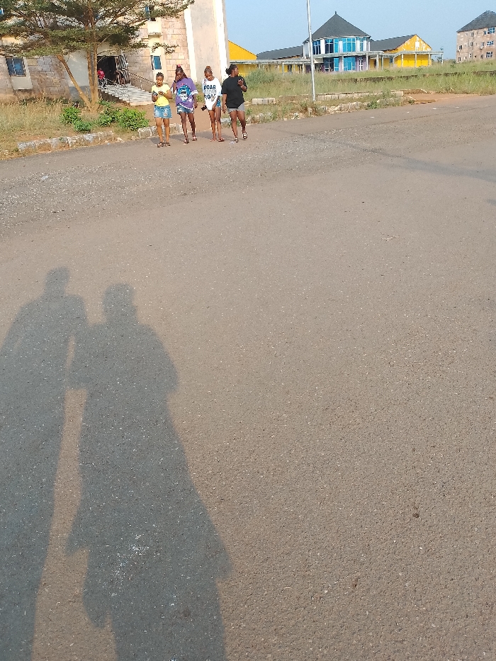 The scene depicts four females walking on a paved asphalt road in Umueshi, Nigeria, under clear daylight, with long shadows cast towards the viewer's right, indicating either early morning or late afternoon.
In the immediate foreground, two distinct, elongated human shadows are present on the asphalt, one to the left and one to the right, suggesting the position of the photographer and an accompanying individual.
In the midground, four females walk in a line from left to right. The leftmost individual wears a light-colored t-shirt and blue shorts, looking down at a mobile phone. The second individual from the left wears a patterned t-shirt and dark shorts, holding an object, possibly a phone or camera, near her chest. The third individual wears a white t-shirt with "ROAR" printed on it and light-colored shorts. The fourth individual on the far right wears a dark t-shirt and dark shorts. All four wear sandals or flip-flops.
The background features multiple structures. To the left, behind the walking individuals, stands a light-colored, possibly stone or concrete, building with a visible entrance and steps. A small tree with green foliage is positioned to the left of this building. Beyond a stretch of dry, unkempt grass, several more buildings are visible: a distinctive structure with a blue, multi-faceted roof and blue-tinted windows, a building with a yellow facade to its right, and a multi-story building further to the right. The sky is clear.