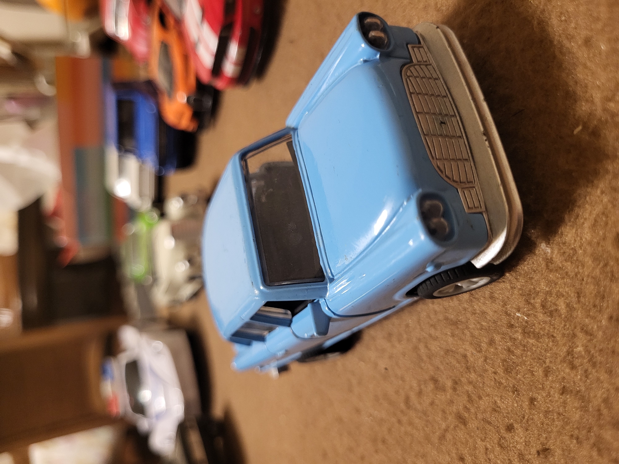 The image shows a blue toy car, possibly a Ford Anglia, on a brown surface. It's surrounded by other toy cars of various colors in the background. The focus is on the blue car, highlighting its details such as the grill and front bumper. The scene appears to be indoors, likely a room with a carpet or fabric surface. There is no information provided that would allow one to determine if the image was taken in Yerevan, Armenia.
