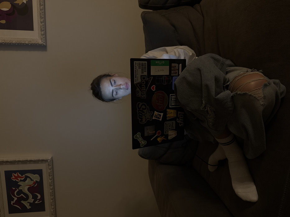 An individual is seated on a dark grey sectional sofa in a dimly lit indoor environment, identified as a location within the city of Venice, Italy. The person, dressed in a white long-sleeved top, ripped light-wash jeans, and white socks with blue stripes, is looking down at a black laptop resting on their lap. The laptop screen emits light, illuminating the person's face. The laptop lid is extensively covered with various stickers, including "Doritos Star," "Lays," "Skoda," a barcode label "PLF4H3M1G," and a "Hello, My Name Is Lena" nametag. The wall behind the sofa is light-colored and features two framed abstract art pieces; one on the left with red, grey, white, and dark blue elements, and a partially visible one on the right with yellow and purple tones.