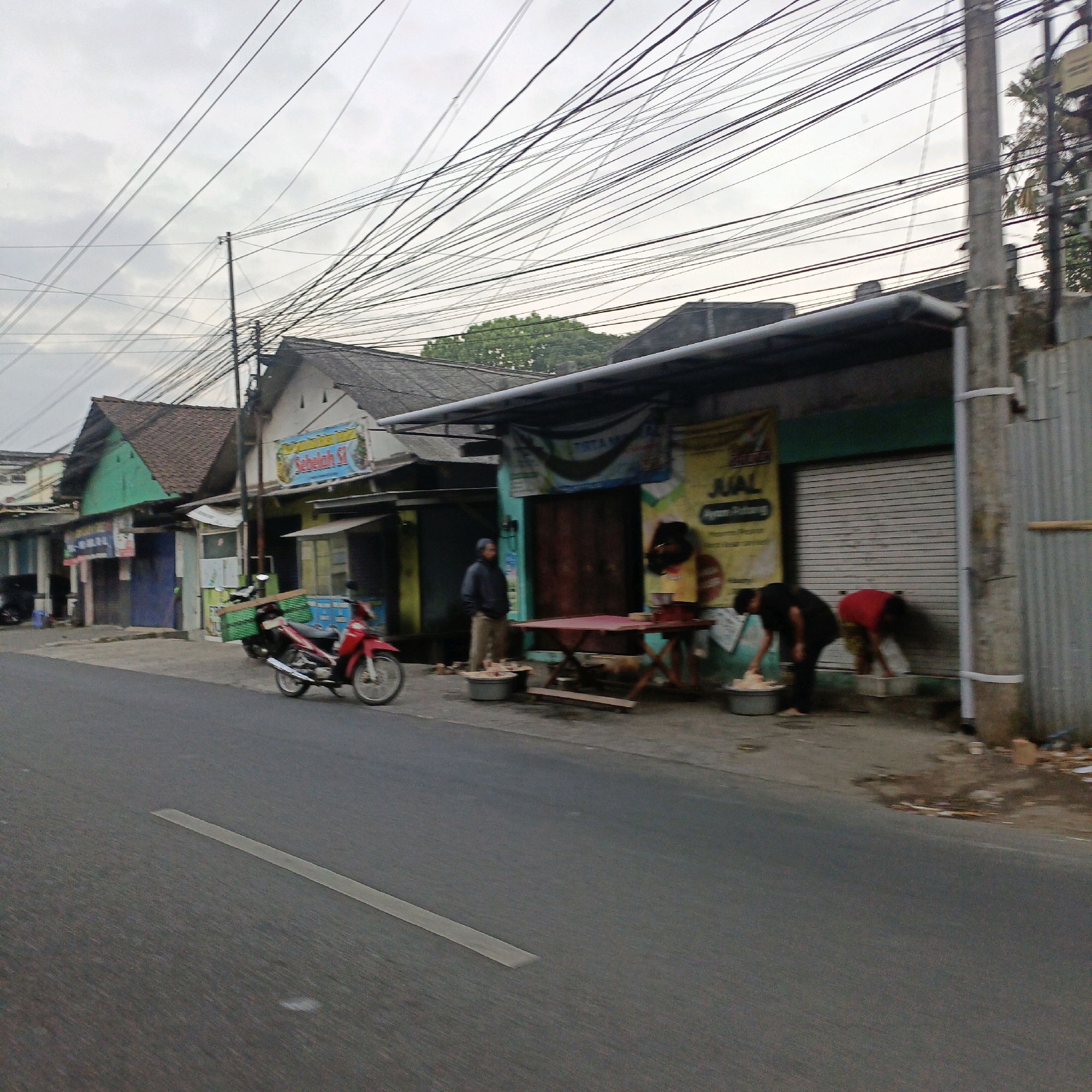 The image depicts a street scene in Salatiga, Indonesia, characterized by a densely packed overhead network of electrical and telecommunication wires. On the right side of the road, a row of modest shopfronts is visible, some with corrugated metal shutters, others with open entrances displaying goods or activities. A prominent sign with Indonesian text, likely advertising a business, is affixed to one of the buildings. In front of the shops, a red motorcycle is parked, and several individuals are engaged in various activities. One man, wearing a blue jacket and beige pants, stands near a large basin. Two other people, one in a black shirt and the other in a red shirt, are bent over, appearing to be washing or preparing something in large basins near the shop entrances. A red folding table is set up on the pavement, and food or other items are displayed on it. The overall impression is of a bustling, informal commercial area typical of many Indonesian cities, with a focus on everyday economic activities and the ubiquitous presence of overhead utility lines.