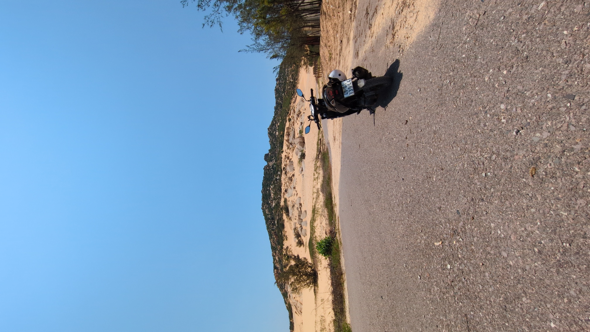 The image shows a motorcycle parked on a paved road in Phuoc Dinh, Vietnam. The motorcycle has a white helmet strapped to the back. A sand dune is located adjacent to the road, with vegetation and rocks visible on the dune. The sky is clear and blue.