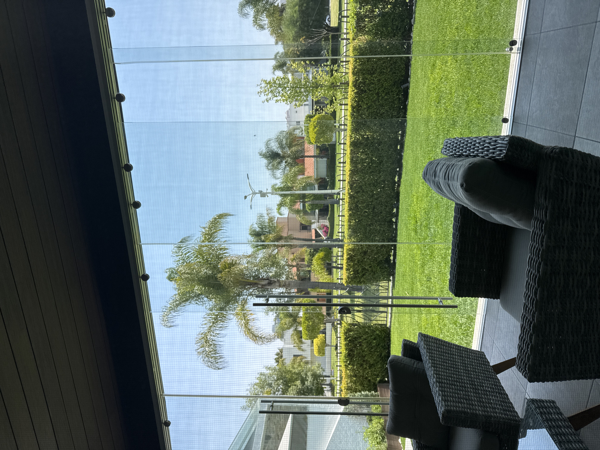 The scene is an indoor patio in San Bernardino Tlaxcalancingo, Mexico, likely during daylight. A grey, cushioned wicker patio set with a table sits on a grey tiled floor. A glass wall or partition with visible metal attachment points separates the patio from the outdoors. The exterior view includes a manicured lawn, tall green hedges, palm trees, other trees and shrubs, buildings, and clear blue sky.